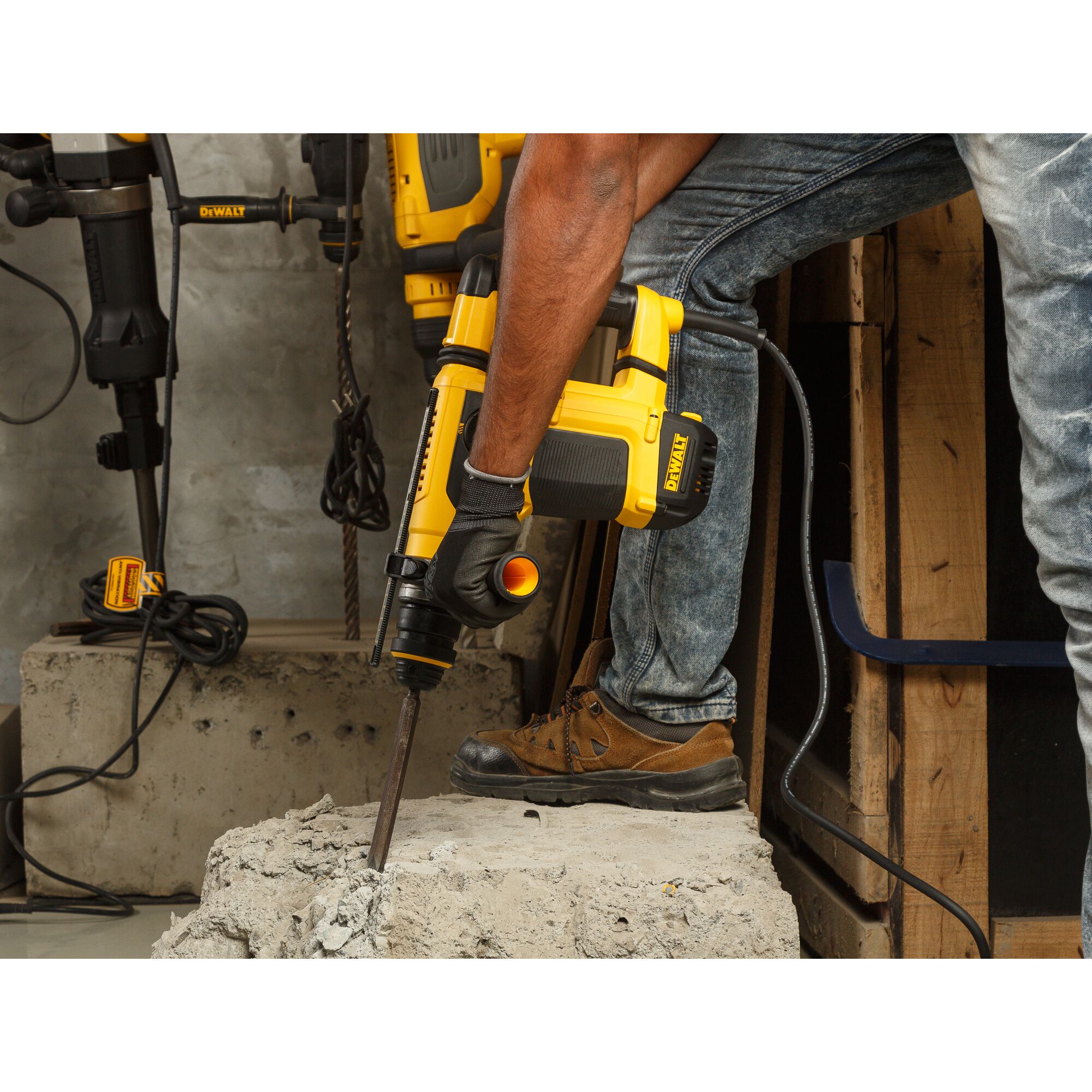 A person using a DEWALT rotary hammer drill (model D25413K) to break concrete in a construction setting. The person is wearing gloves, jeans, and brown work boots. The tool is plugged in and other power tools are visible in the background.