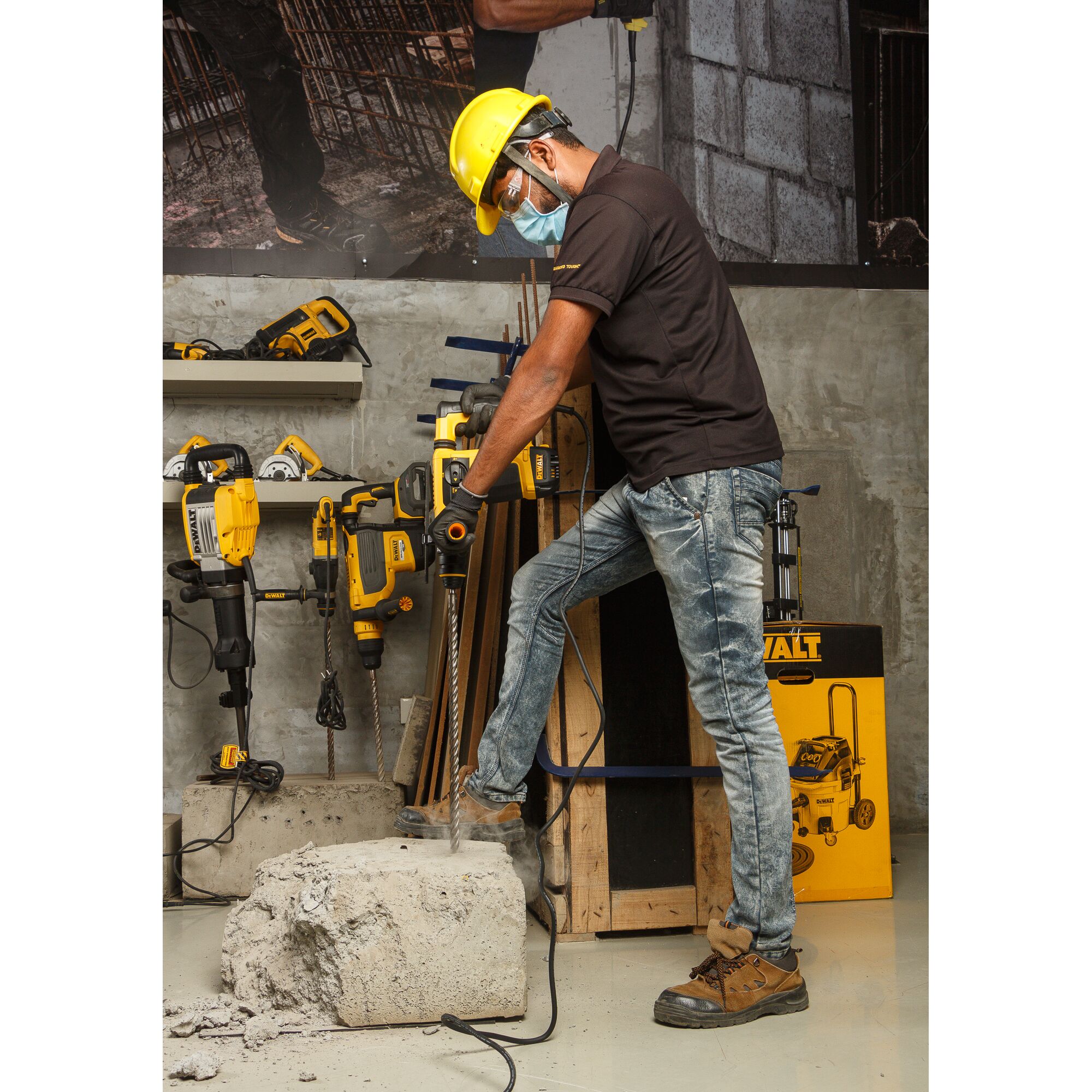 A person wearing safety gear uses a DEWALT hammer drill to break a large concrete block in a workshop. Other DEWALT power tools are visible in the background, along with construction materials.