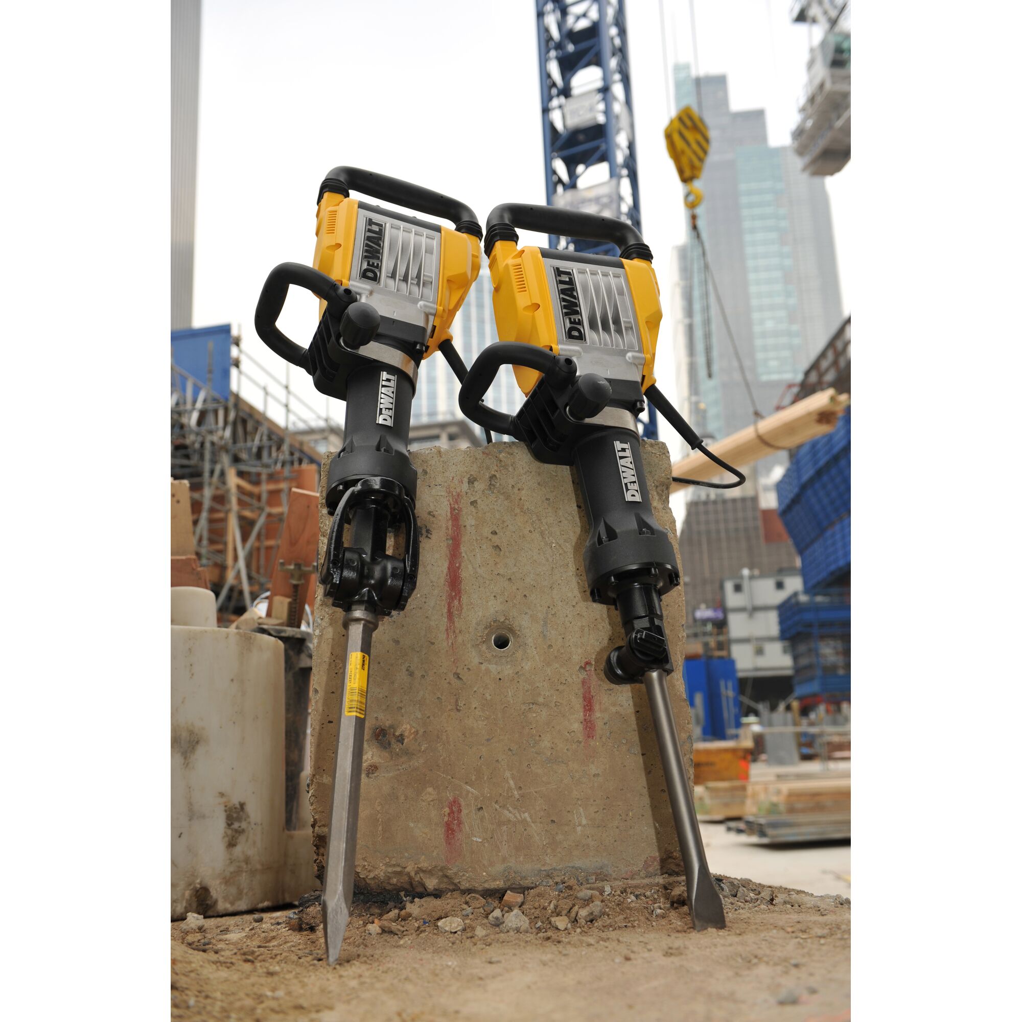 Two DEWALT electric demolition hammers with yellow and black design are positioned upright on a concrete slab at a construction site, with cranes and high-rise buildings visible in the background.