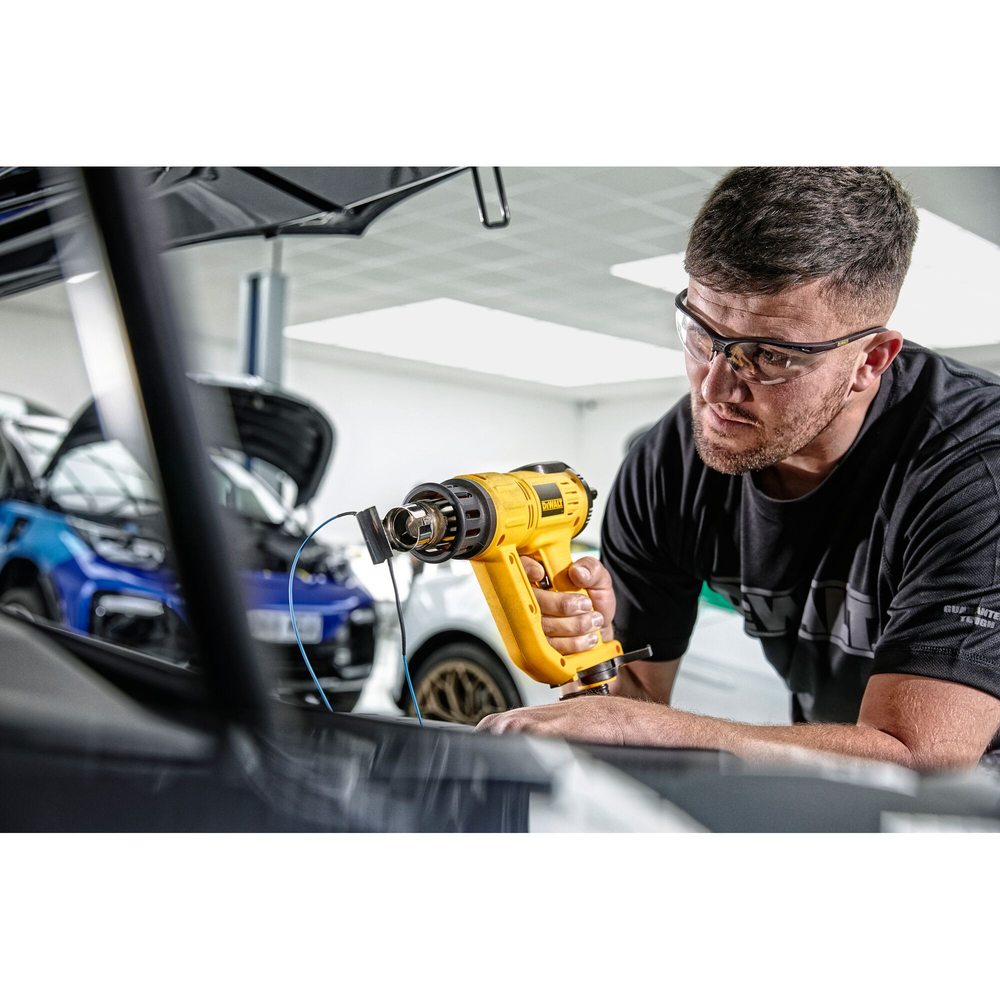 A person using a yellow DEWALT heat gun (model D26414) inside a car workshop. The person's face is blurred, and there are cars with open hoods in the background.