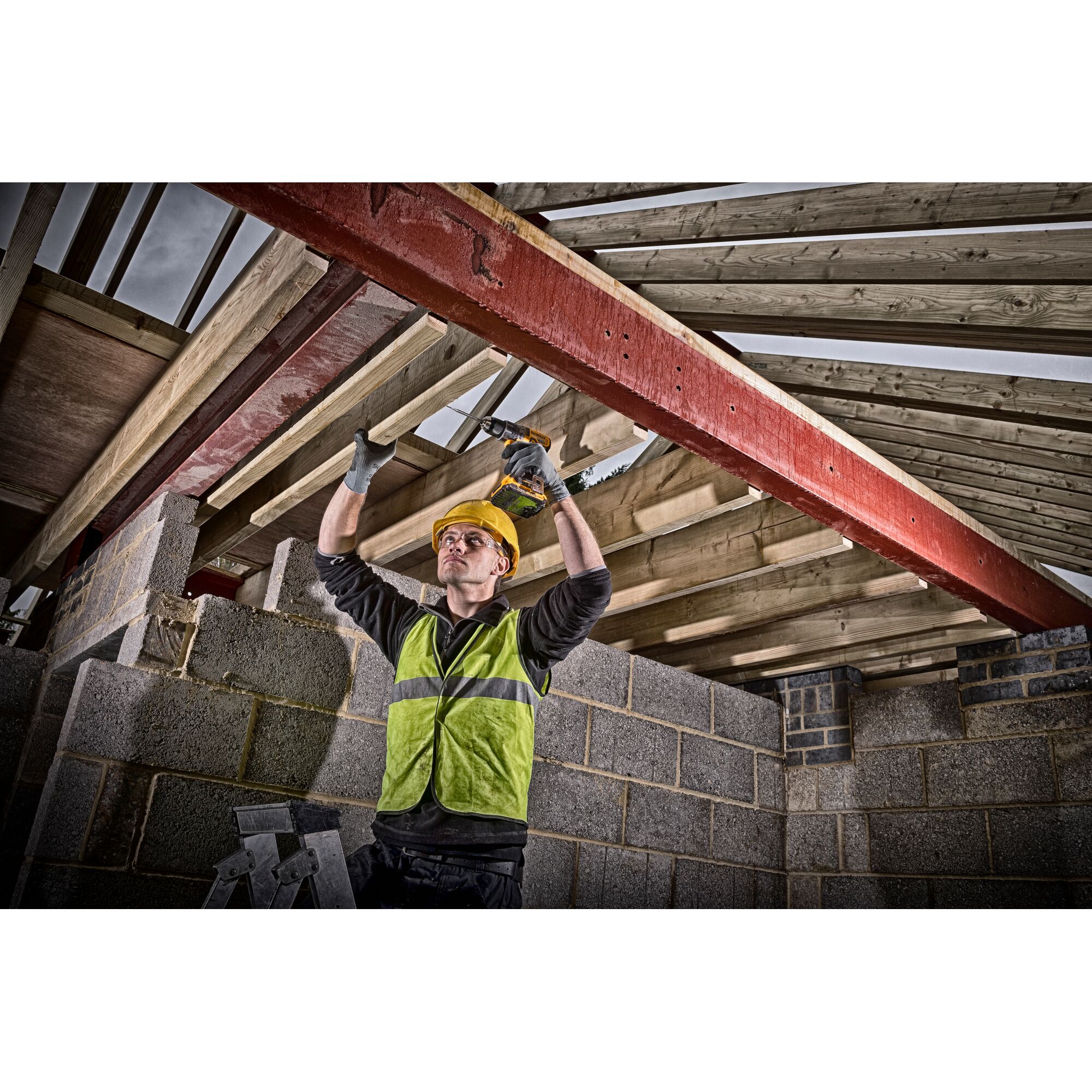 A construction worker wearing a yellow hard hat and a green high-visibility vest uses a power drill on wooden beams in a partially built structure with grey brick walls.