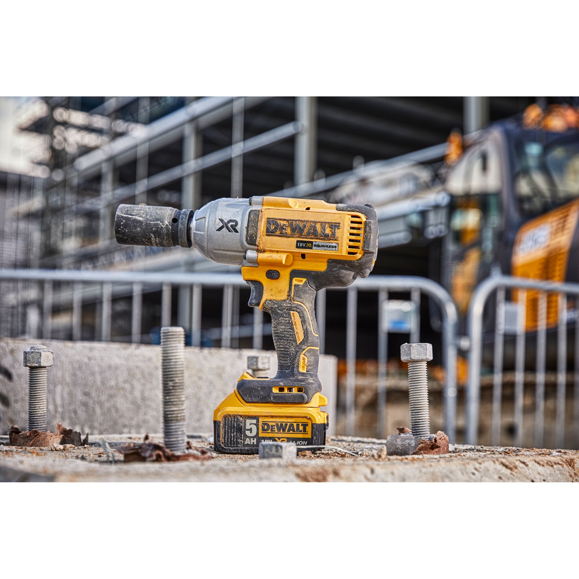 A DEWALT cordless impact wrench is shown standing upright on a construction site, surrounded by large bolts and concrete debris. Industrial equipment and a metal fence are visible in the background.