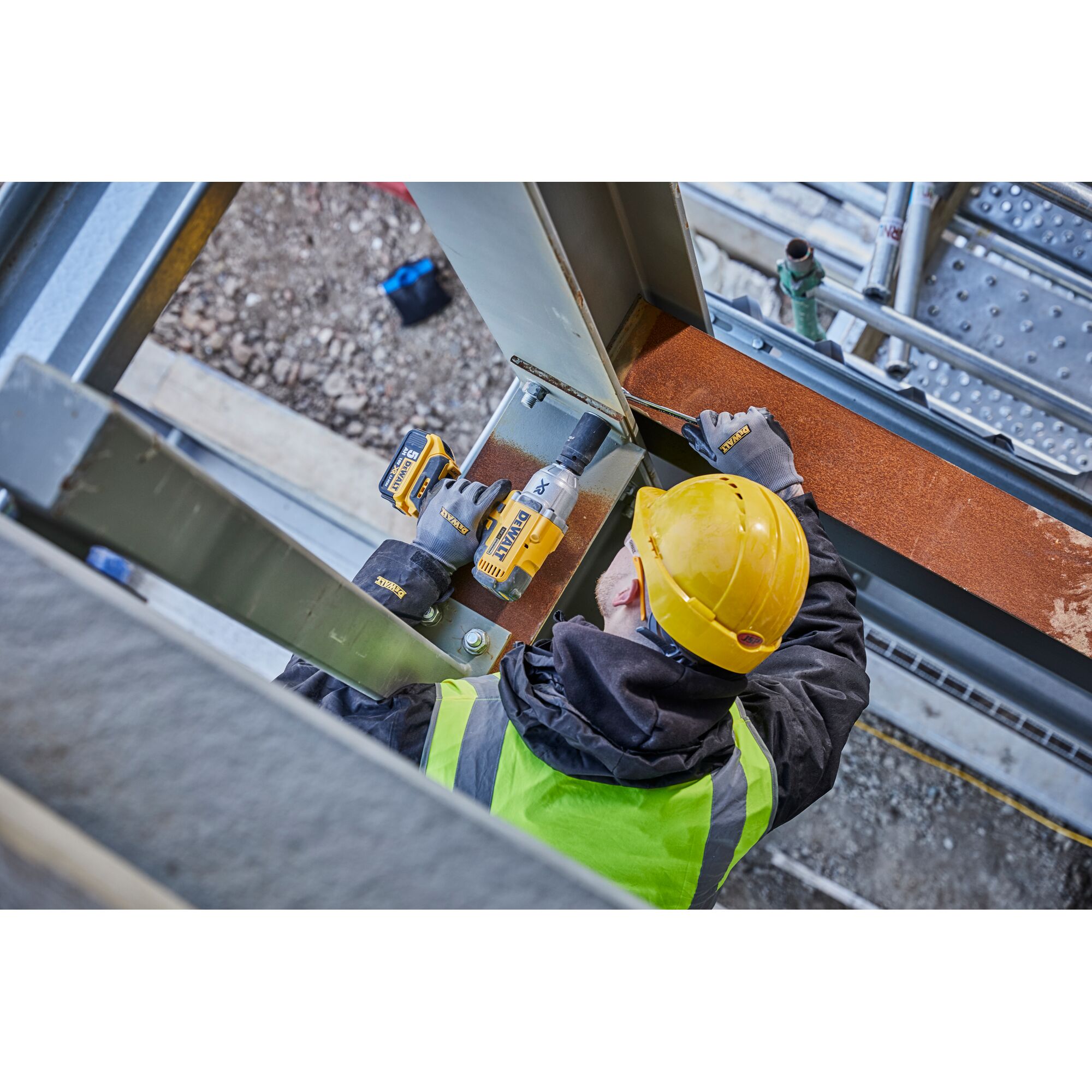 A person wearing a yellow hard hat and high-visibility vest uses a DEWALT cordless impact wrench to fasten bolts on a steel beam at a construction site.