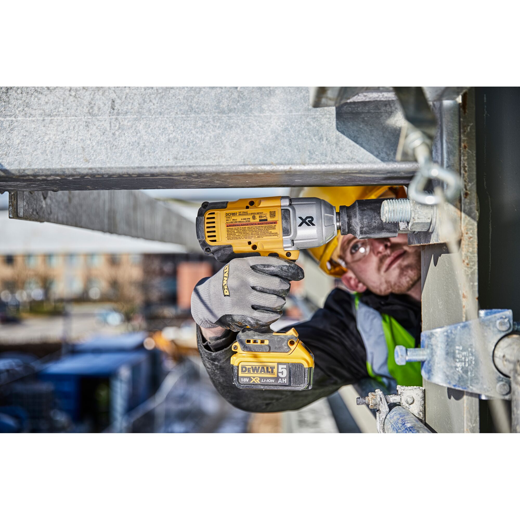 A person wearing protective gloves and a helmet uses a DEWALT cordless impact wrench (model DCF897N) to fasten a large bolt on a steel structure at a construction site.
