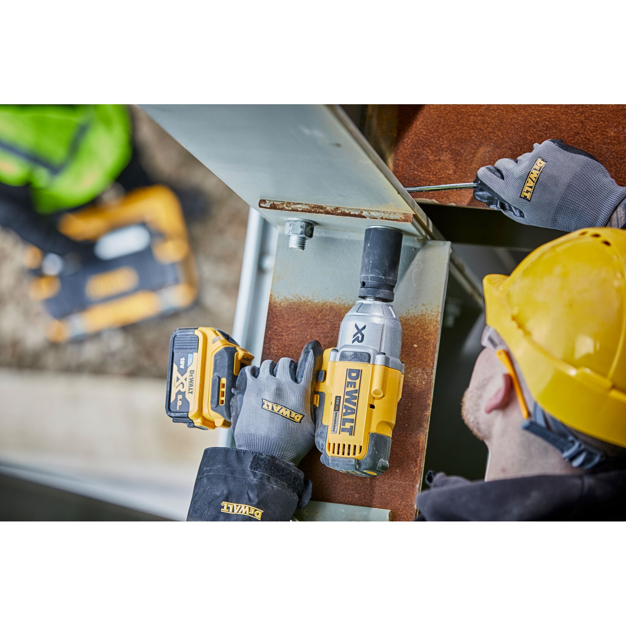 A worker wearing a yellow hard hat and DEWALT gloves uses a DEWALT cordless impact wrench (model DCF897N) on a steel beam at a construction site.