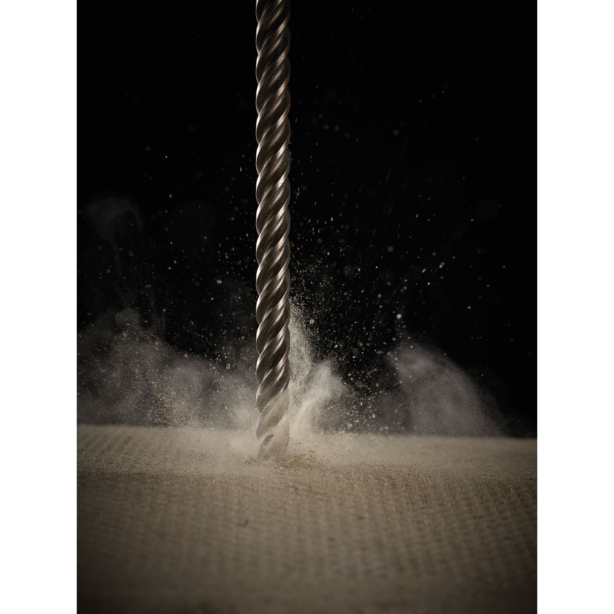 Close-up view of a metal drill bit cutting through a rough surface, with dust and particles scattering around against a dark background.