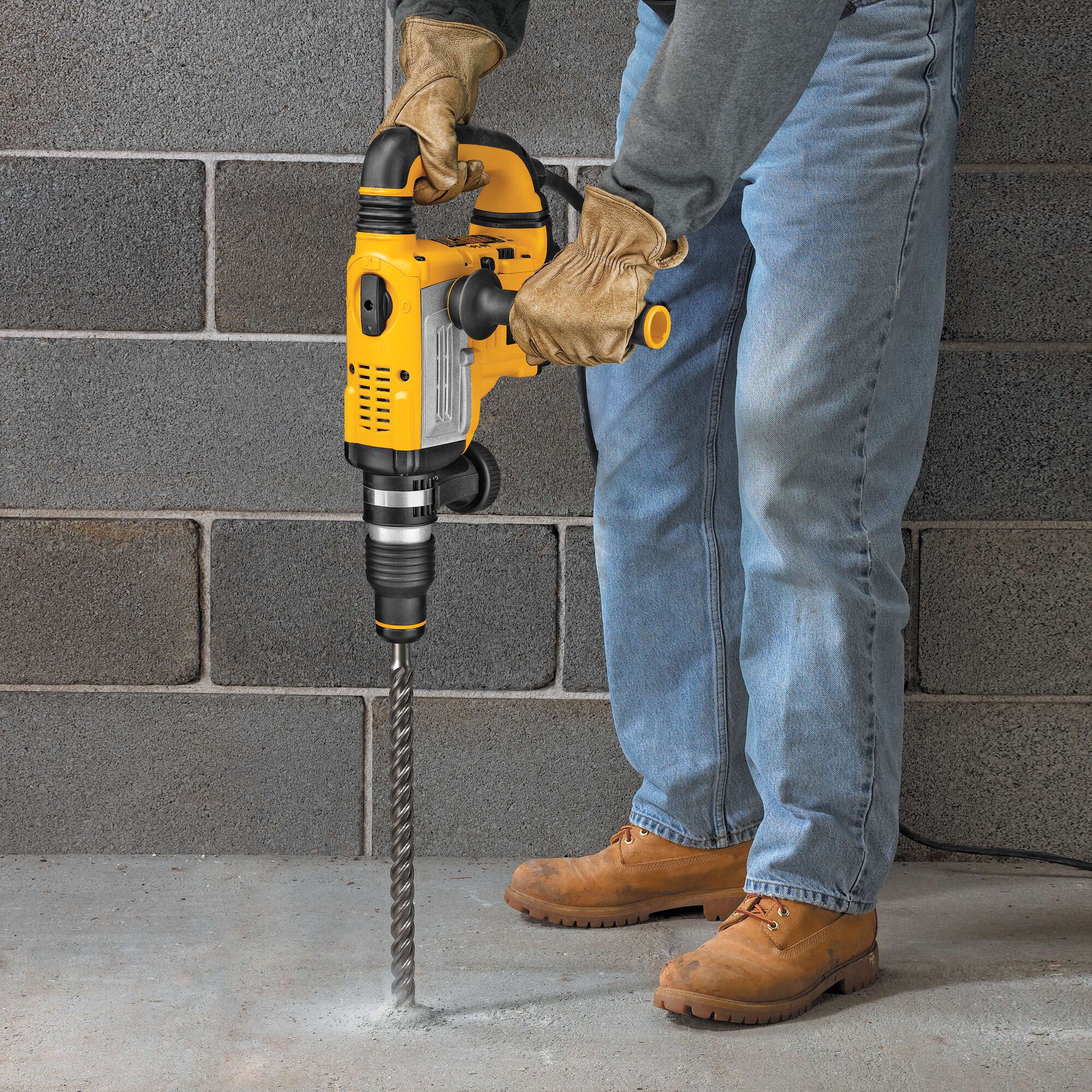 A person wearing gloves and work boots is using a yellow DEWALT rotary hammer drill with a long drill bit to drill into a concrete floor. The person is standing in front of a gray concrete block wall.