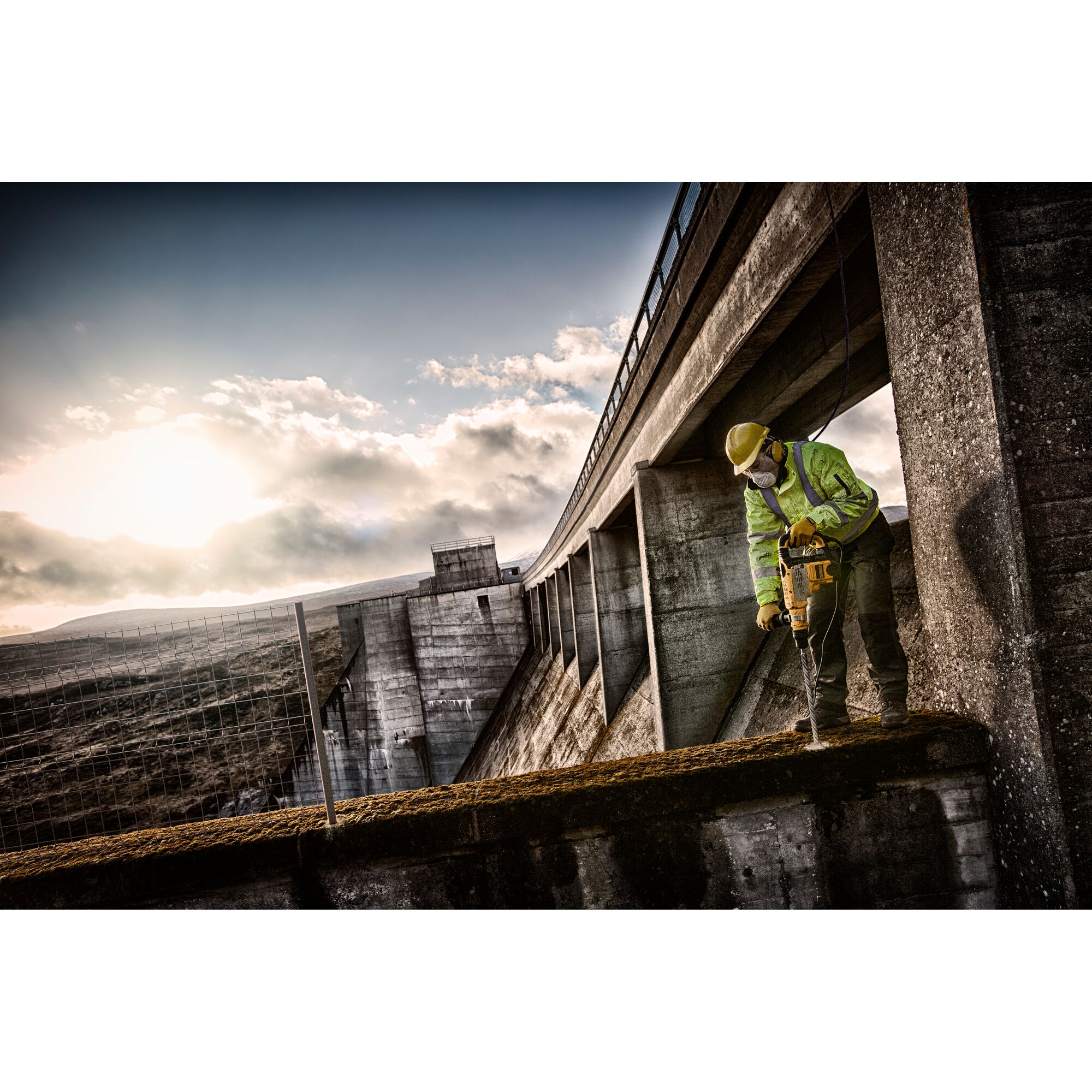 A construction worker wearing safety gear uses a DEWALT power tool to drill into concrete on a large industrial structure, possibly a dam, during sunrise or sunset.