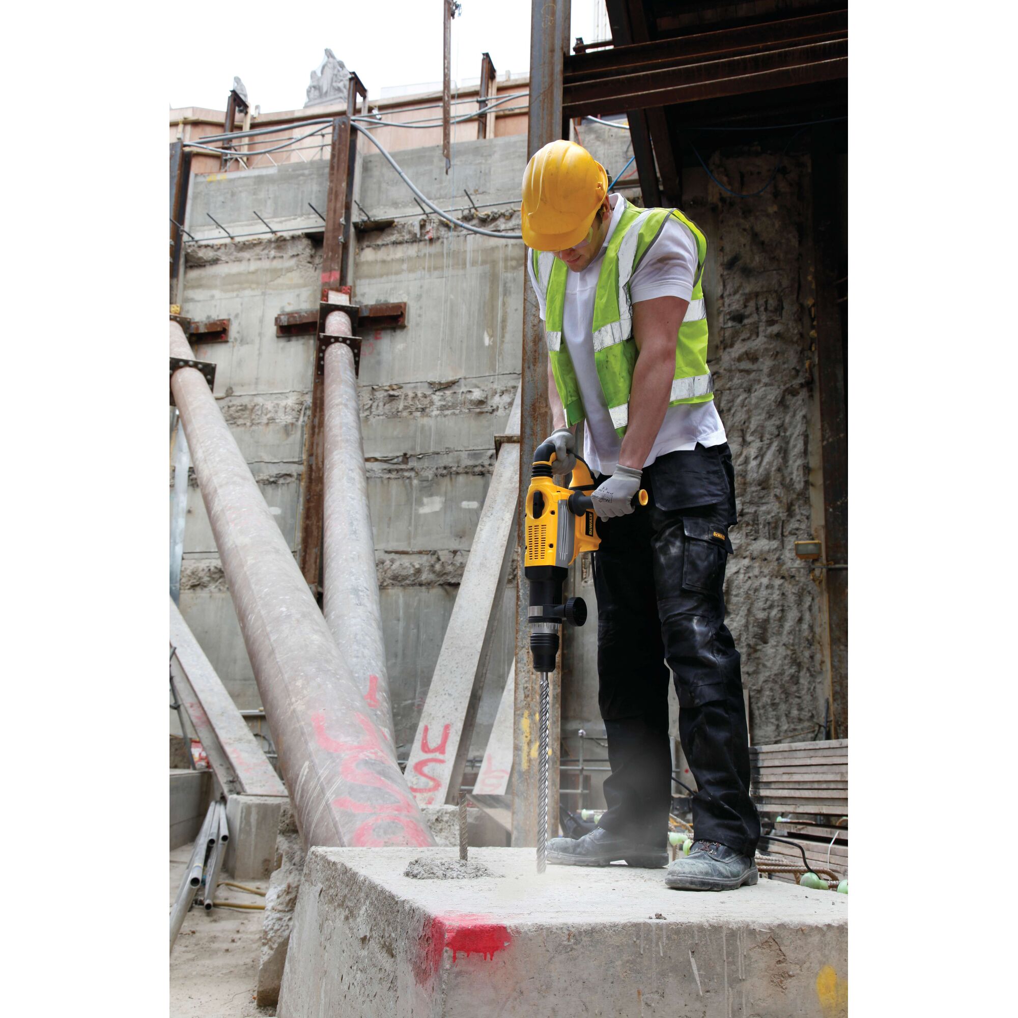 A construction worker wearing a yellow hard hat and reflective vest is using a DEWALT power tool to drill into a concrete block at an industrial construction site.