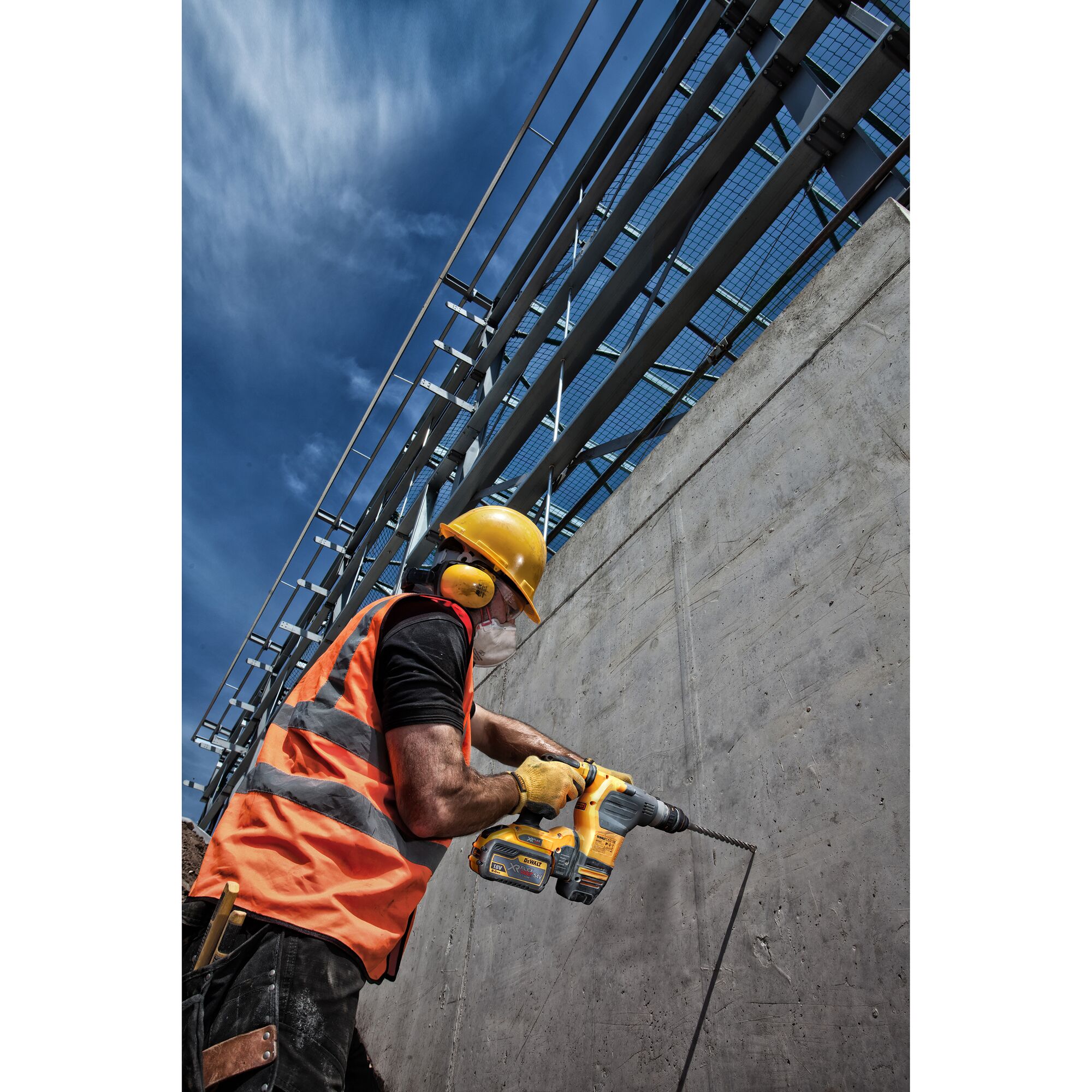 A construction worker wearing a yellow hard hat, orange safety vest, ear protection, gloves, and a mask is using a yellow DEWALT power drill to make a hole in a concrete wall at a building site under a blue sky.