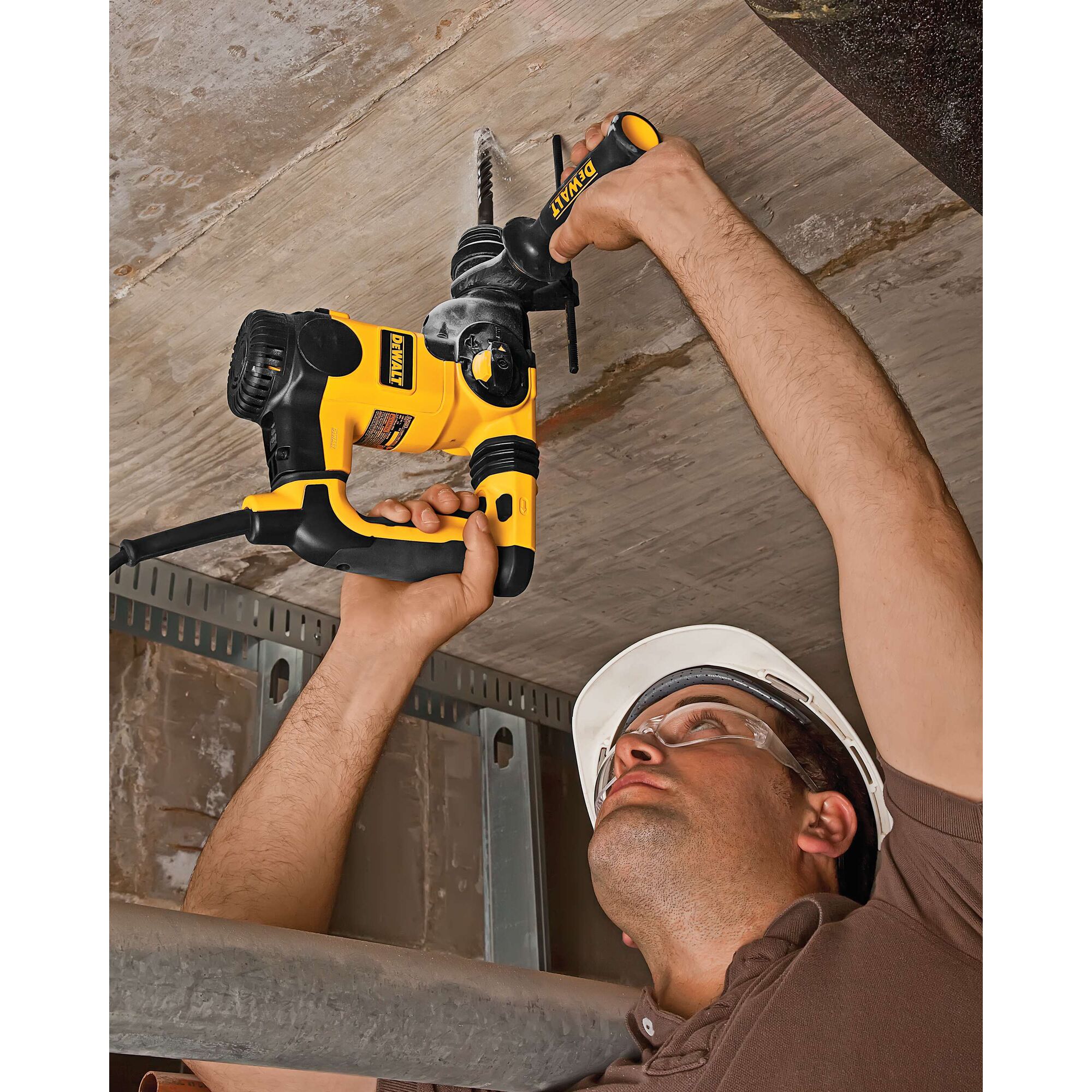 A person wearing a white safety helmet is using a yellow and black DEWALT rotary hammer drill to drill into a concrete ceiling. The person's face is blurred for privacy.