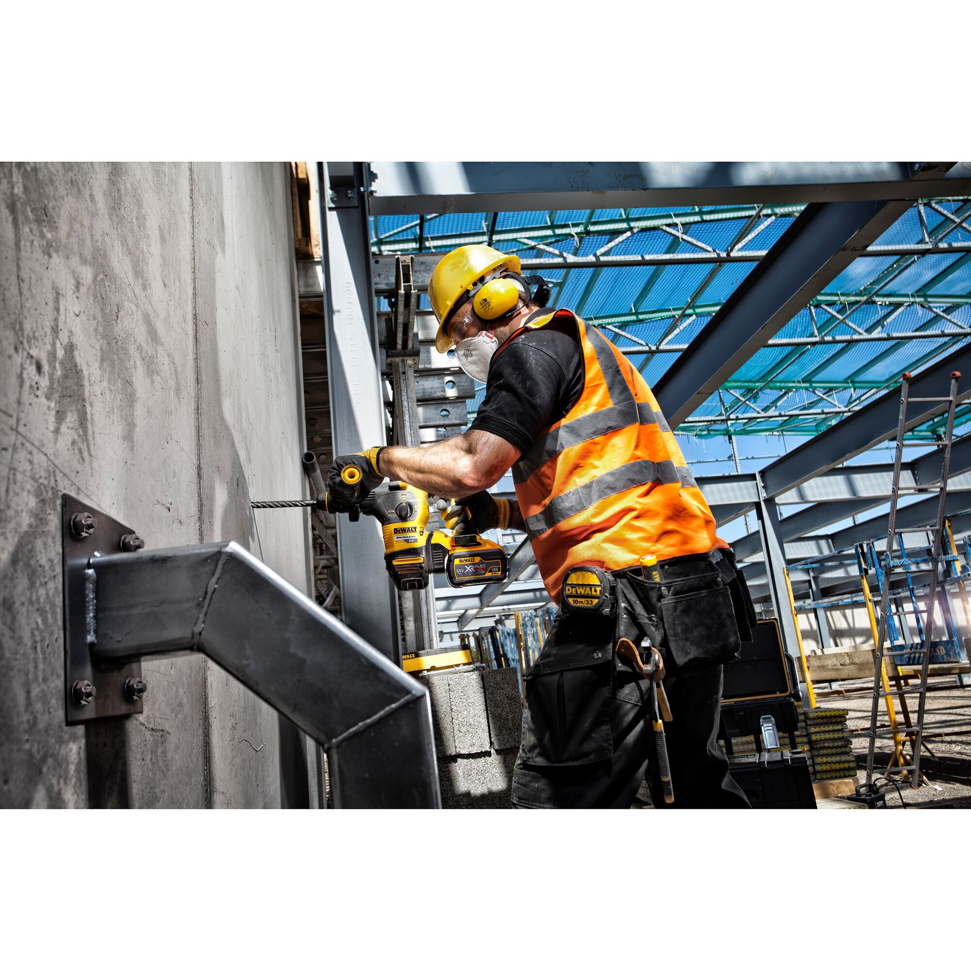 A construction worker wearing a yellow hard hat, ear protection, and an orange safety vest uses a DEWALT power drill on a concrete wall at an industrial site. The worker is equipped with safety gear and tools, and the area contains steel beams and construction materials.