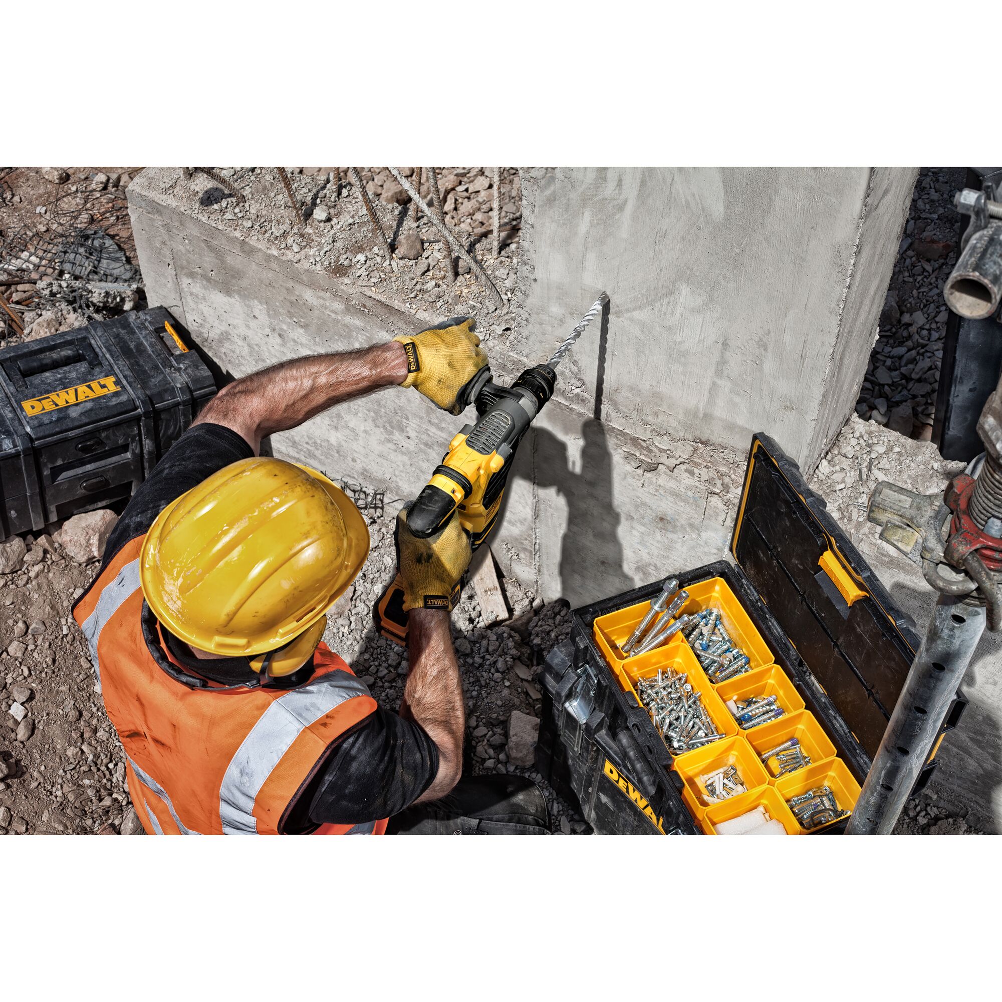 A construction worker wearing a yellow hard hat and orange safety vest is using a DEWALT rotary hammer drill to bore into a concrete wall. There are DEWALT toolboxes nearby, one open with assorted screws and anchors.
