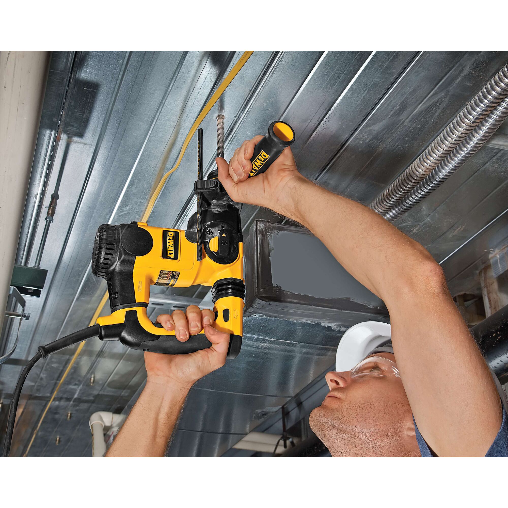 A person uses a DEWALT rotary hammer drill (model DT9549) to drill into a metal ceiling, holding the yellow and black tool with both hands. The person's face is blurred.