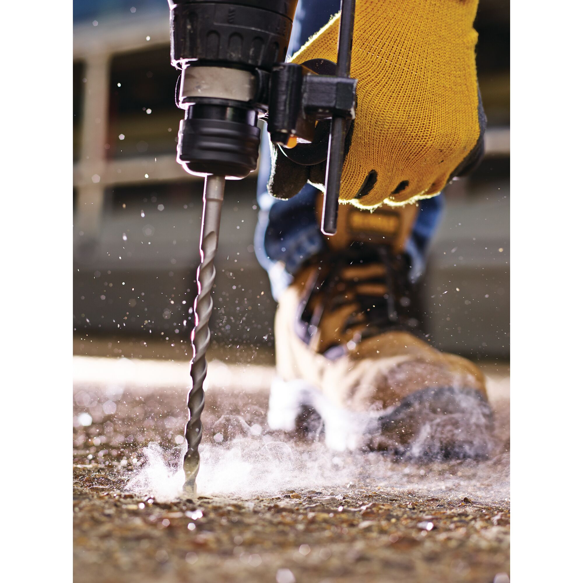 Close-up of a gloved hand operating a power drill with a spiral drill bit on a concrete surface, with dust and debris scattering.