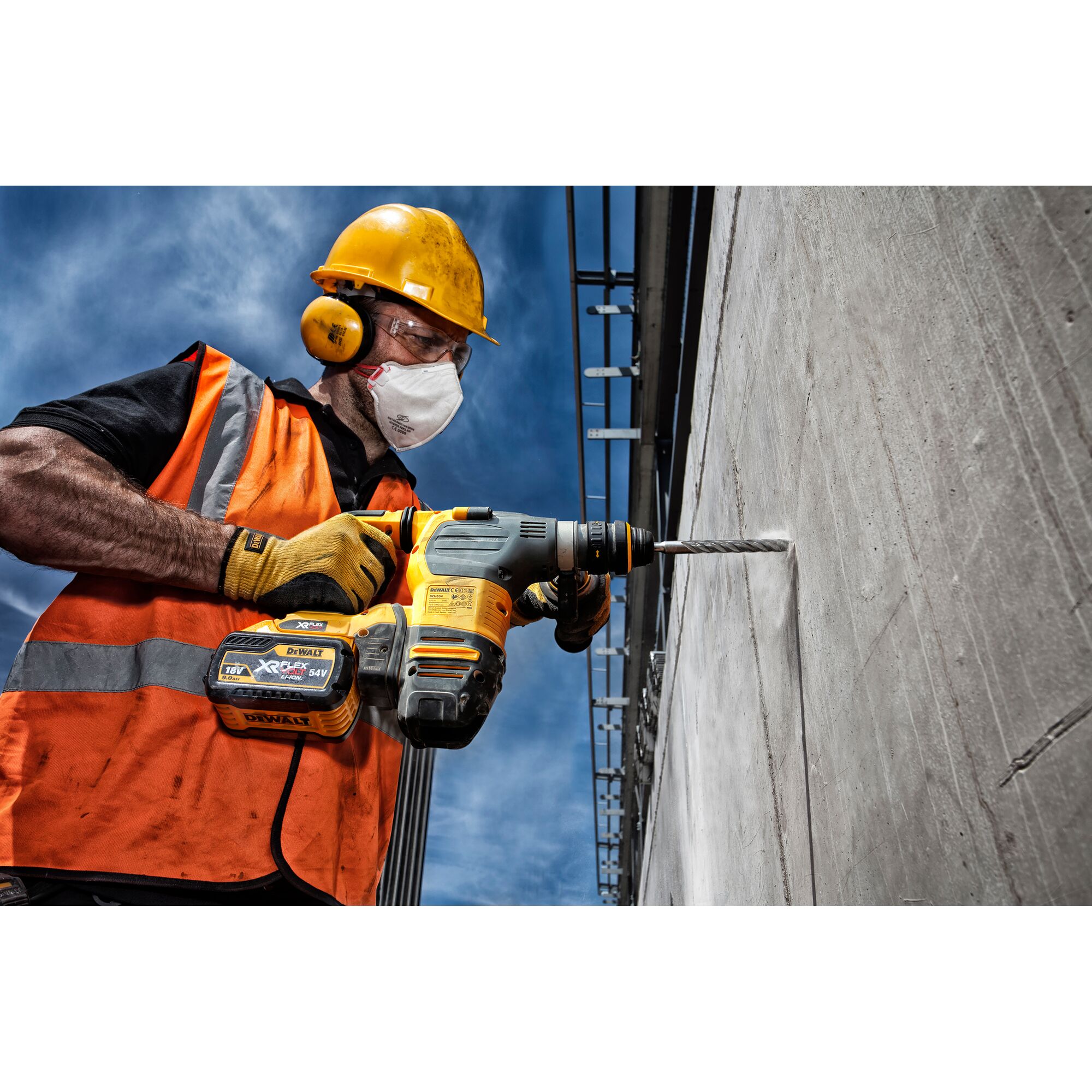 A construction worker in a safety helmet and high-visibility vest is using a DEWALT cordless rotary hammer drill to drill into a concrete wall.
