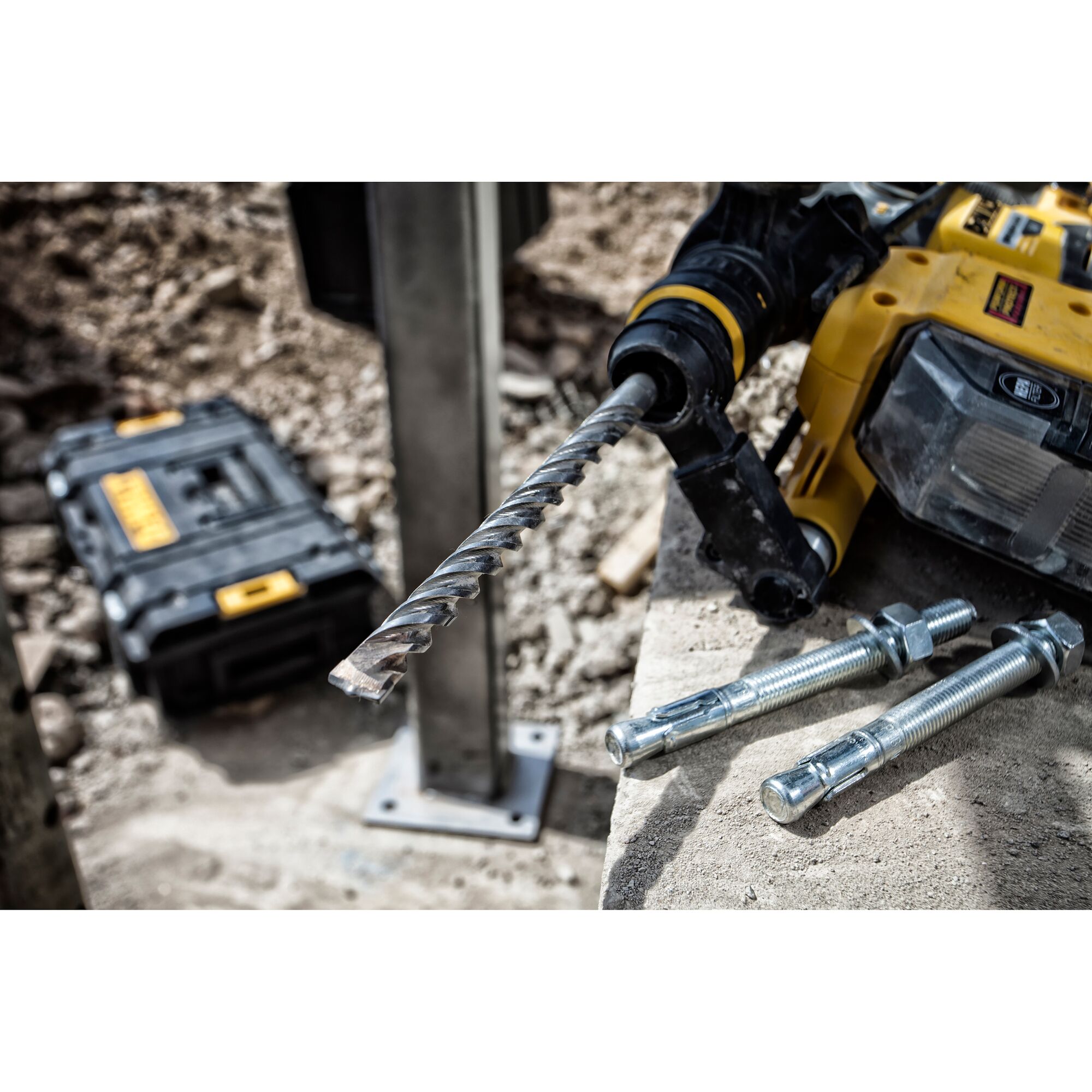 Close-up of a DEWALT power drill with a spiral drill bit, placed on a concrete surface next to three large anchor bolts. A DEWALT tool case is visible in the background at a construction site.