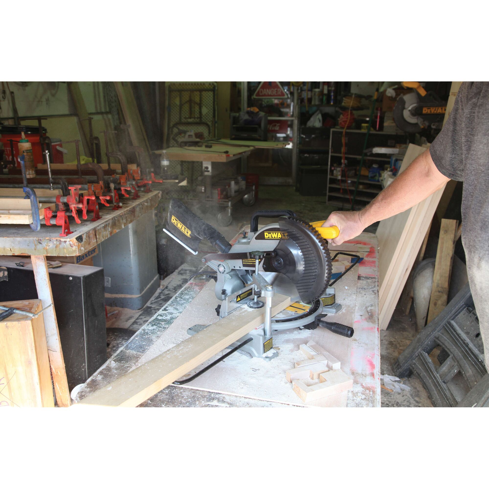 A person operating a DEWALT miter saw, model DW714, to cut a piece of wood in a workshop setting. Sawdust and woodworking tools are visible on and around the workbench.