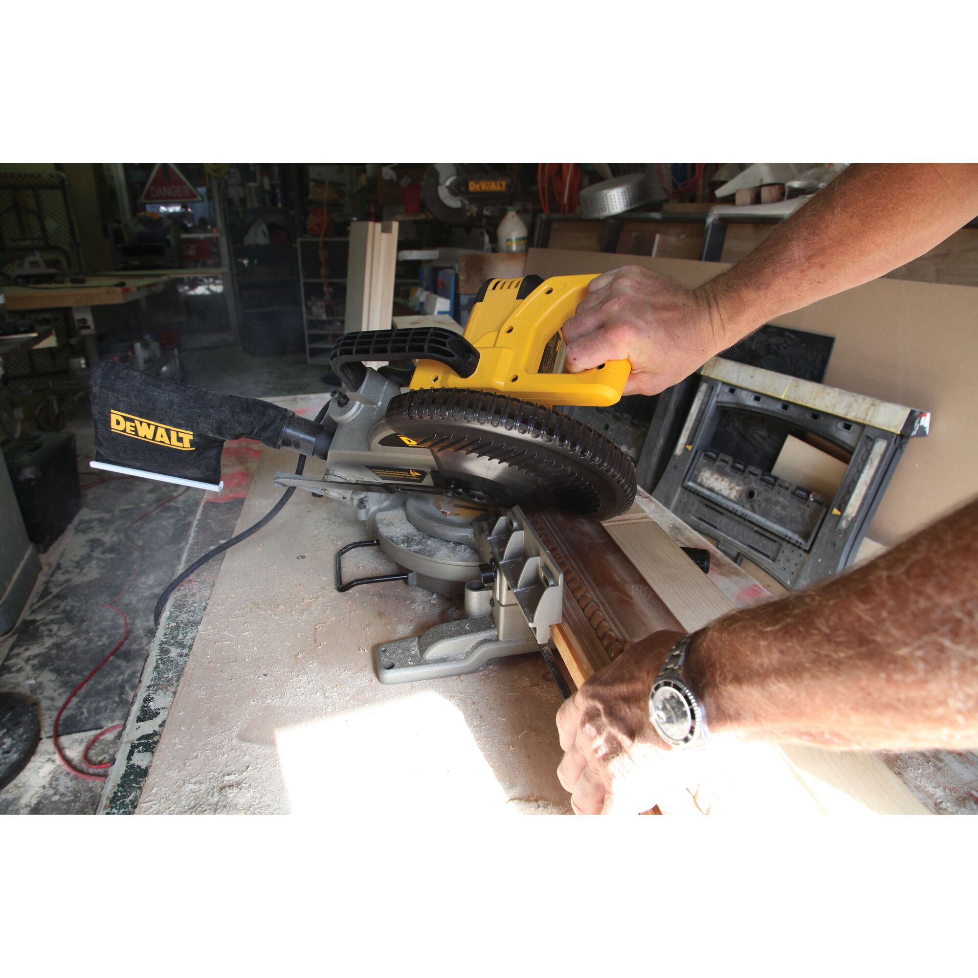 A person is using a DEWALT DW714 miter saw to cut a piece of wood in a workshop. The saw is yellow and black, and there is sawdust on the workbench. The person's hands are holding the wood and operating the saw.