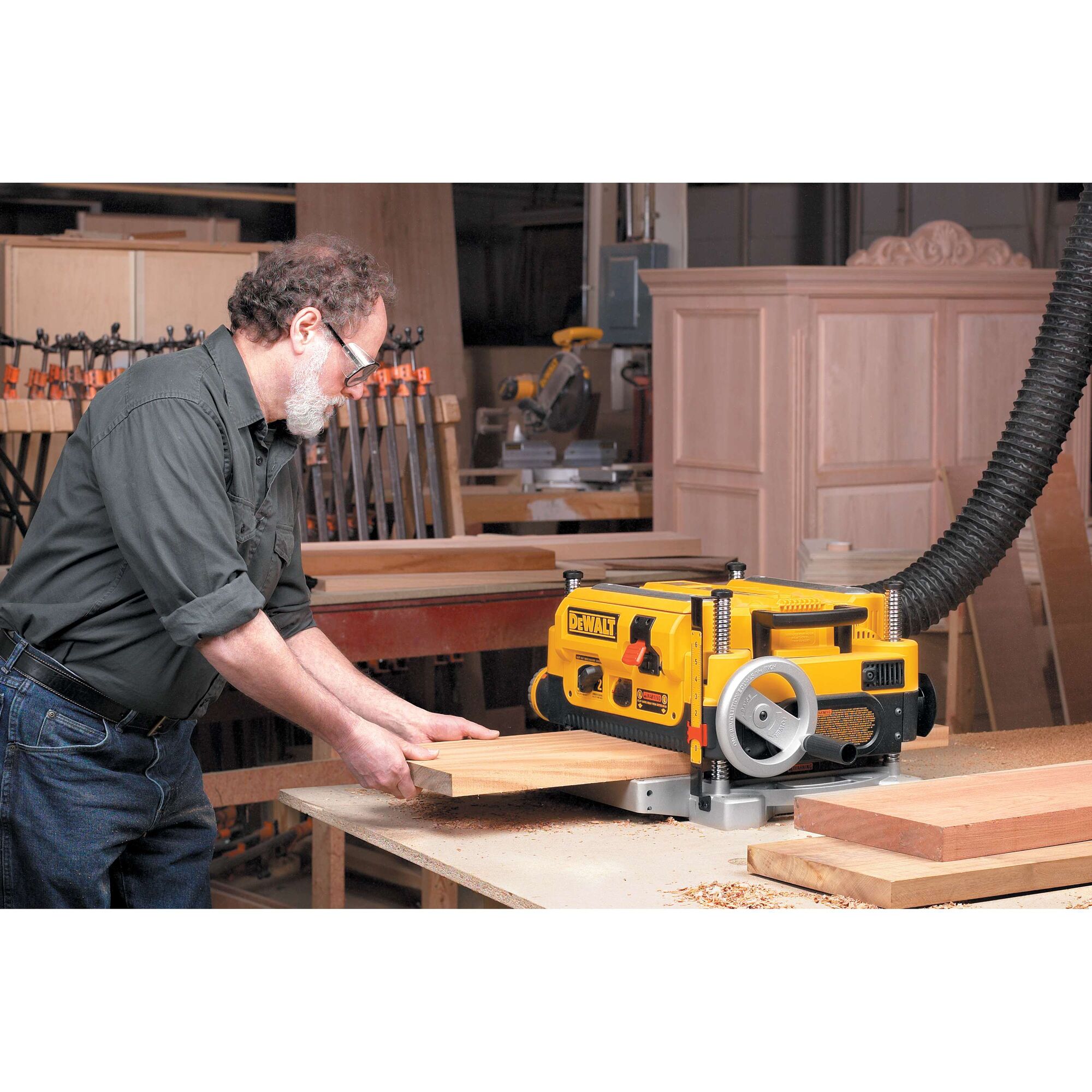 A person using a DEWALT DW735 thickness planer in a woodworking shop, feeding a wooden board through the machine. Various woodworking tools and materials are visible in the background.