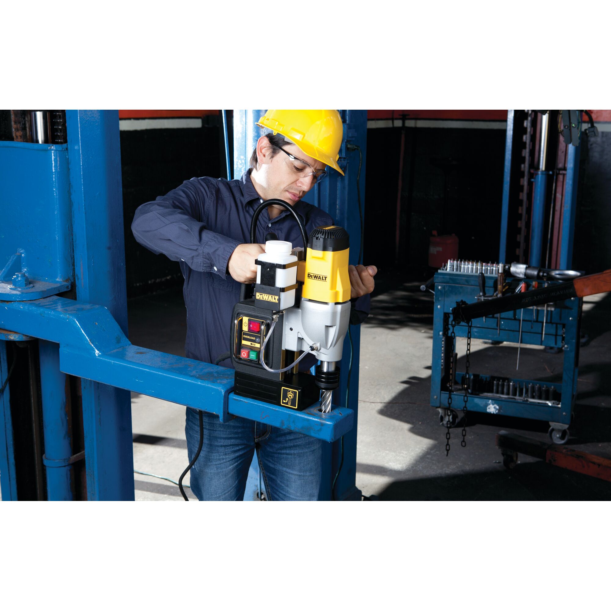 A person wearing a yellow hard hat is operating a DEWALT DWE1622K magnetic drill press on a blue metal structure in an industrial setting.
