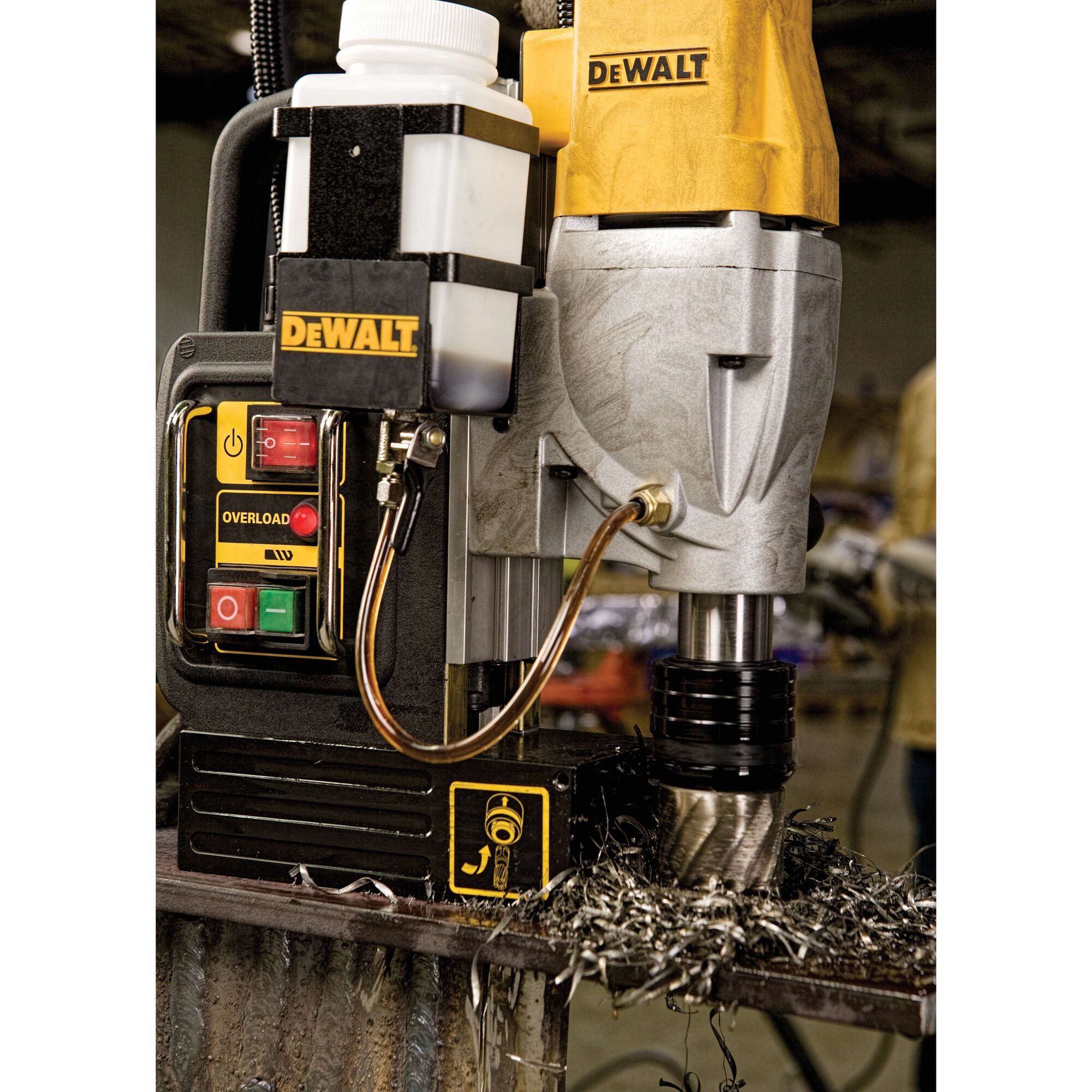 A close-up view of a DEWALT DWE1622K magnetic drill press in use, showing its control panel, coolant bottle, and drill bit cutting through metal with metal shavings around the base.