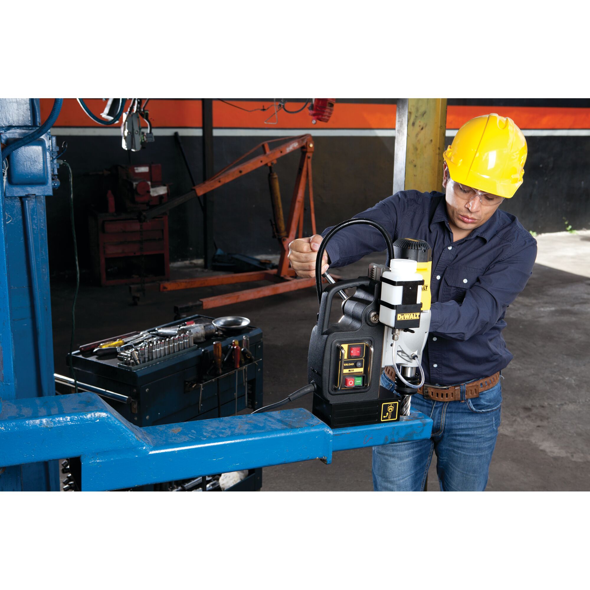 A person wearing a yellow safety helmet operates a DEWALT DWE1622K magnetic drill press on a blue metal beam in an industrial workshop, with a toolbox and various tools visible nearby.