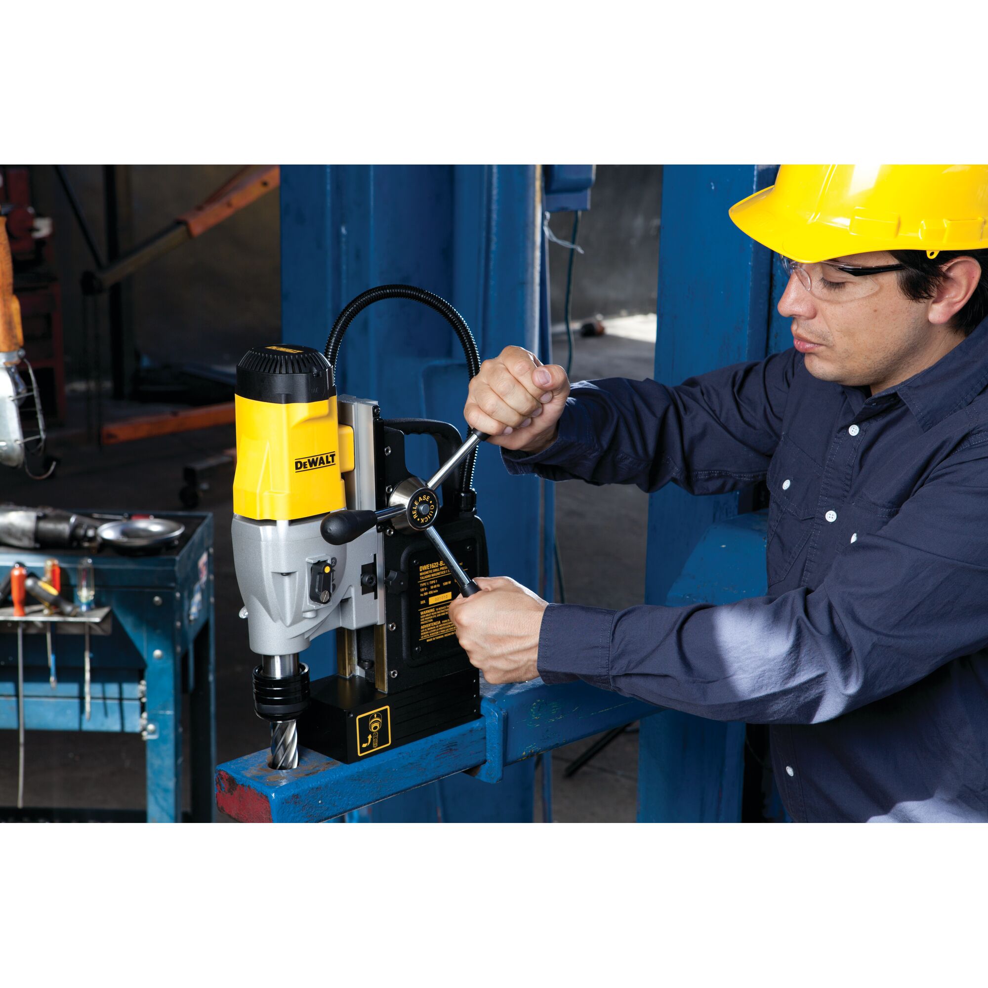 A person wearing a yellow hard hat and blue shirt operates a DEWALT DWE1622K magnetic drill press on a metal beam in an industrial workshop. The machine is clamped to the beam and various tools are visible on a nearby table. The person's face is blurred for privacy.