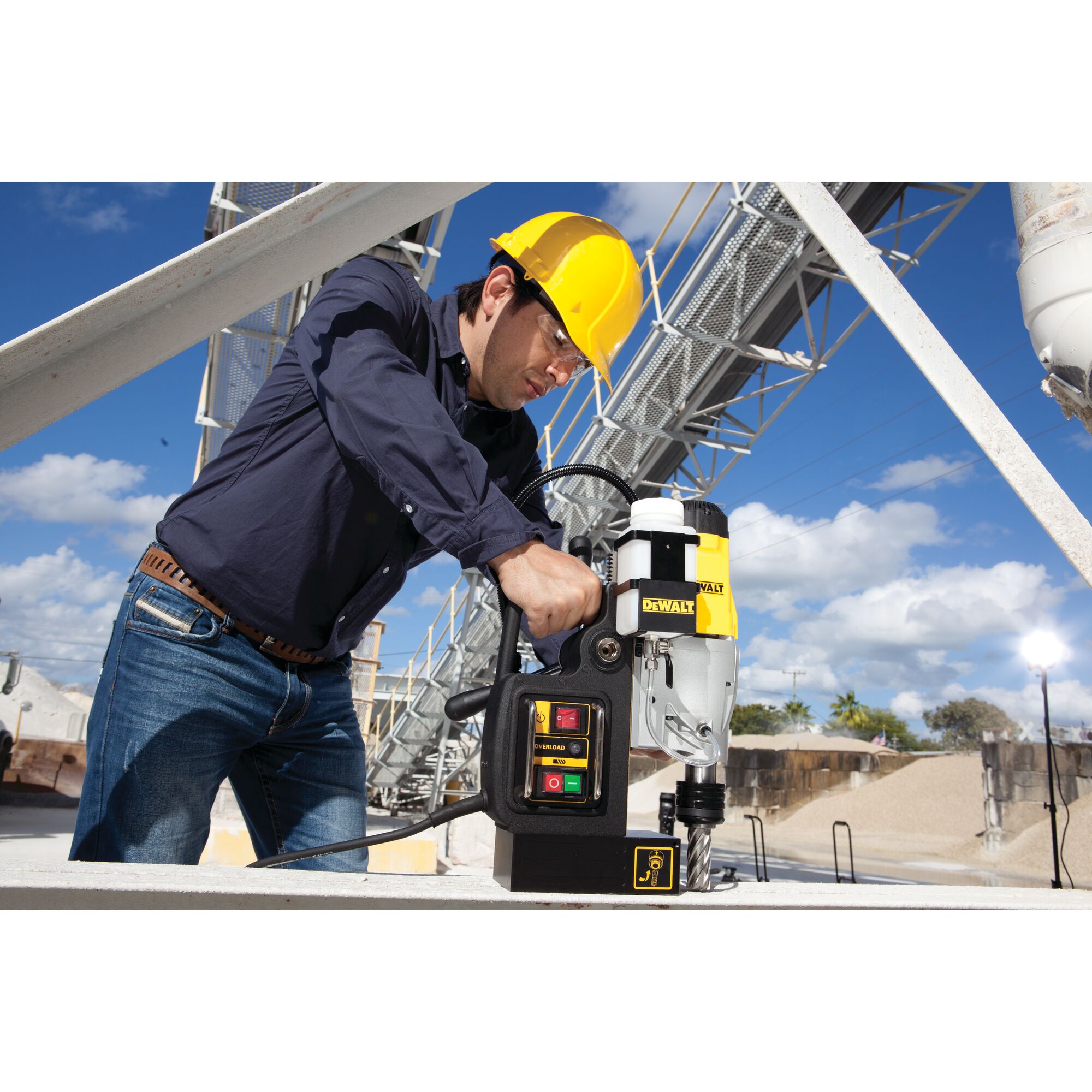 A person wearing a yellow hard hat operates a DEWALT DWE1622K magnetic drill press on a metal surface outdoors, with industrial structures and a blue sky in the background.