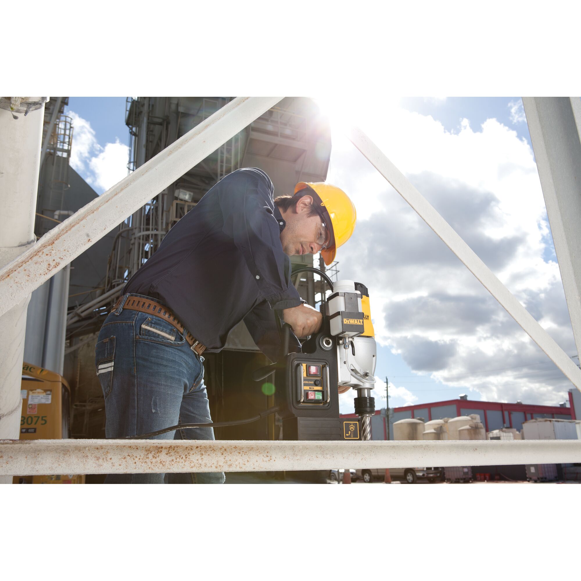 A person wearing a yellow safety helmet and dark clothing is operating a DEWALT DWE1622K magnetic drill press on a metal structure at an industrial outdoor site. The scene is well lit with sunlight and includes construction equipment and buildings in the background.