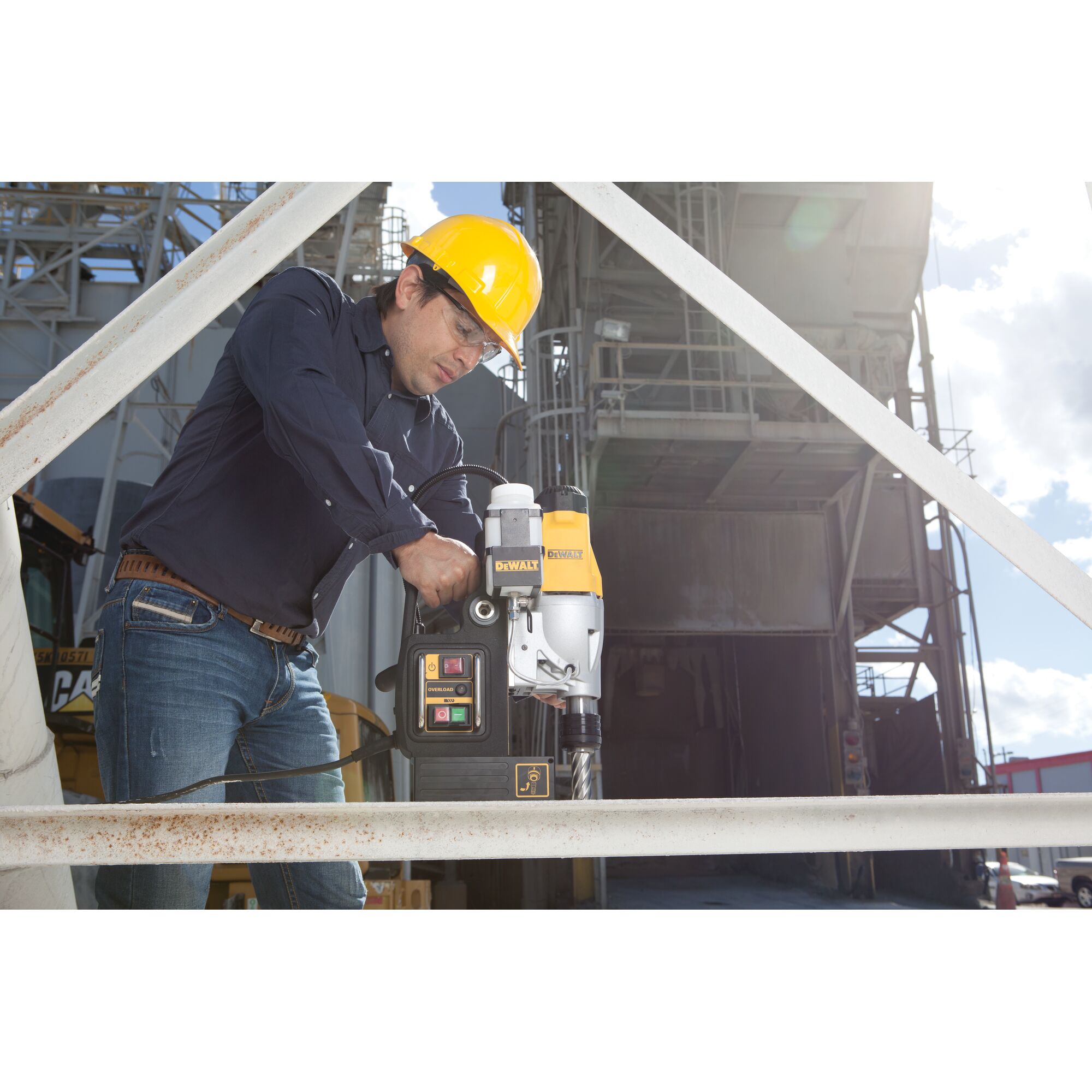 A person wearing a yellow safety helmet operates a DEWALT DWE1622K magnetic drill press on a metal beam at an industrial work site. The person's face is blurred for privacy.