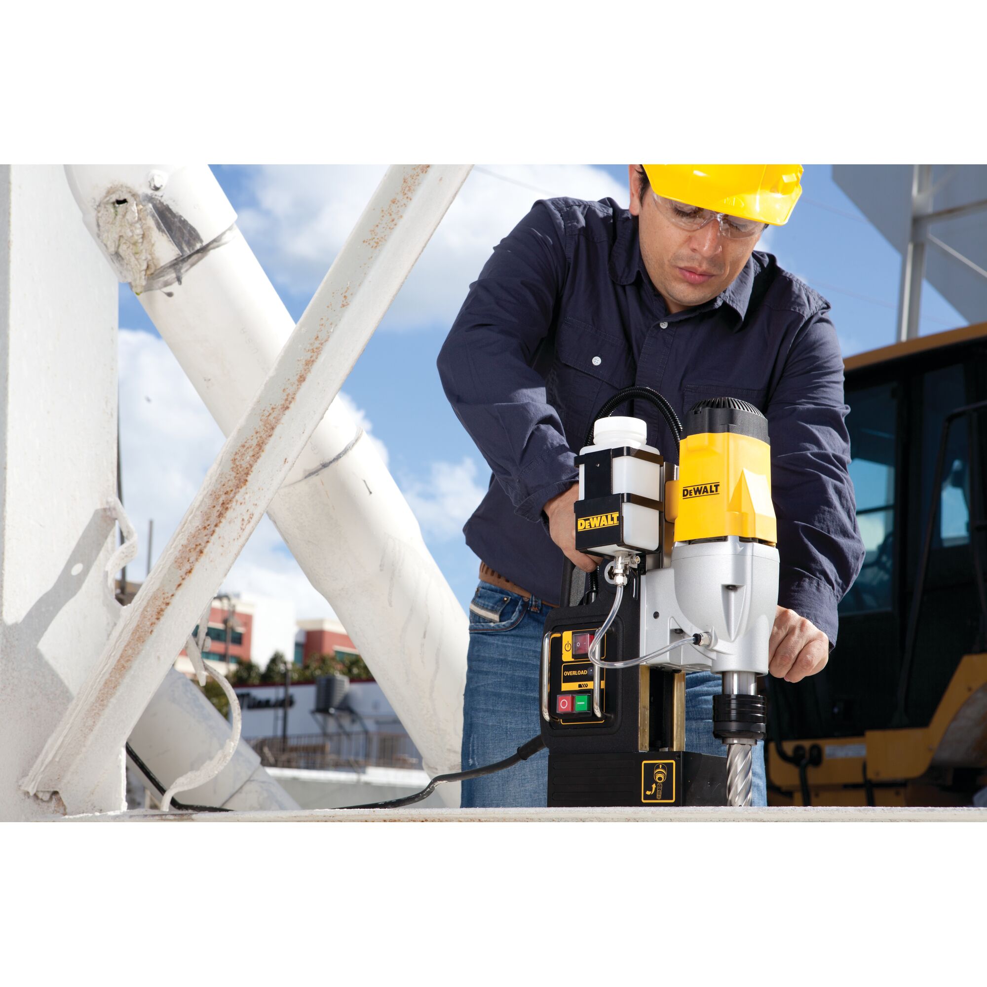 A person using a DEWALT DWE1622K magnetic drill press on a metal surface at a construction site. The person’s face is blurred and they are wearing a blue shirt and yellow safety helmet.