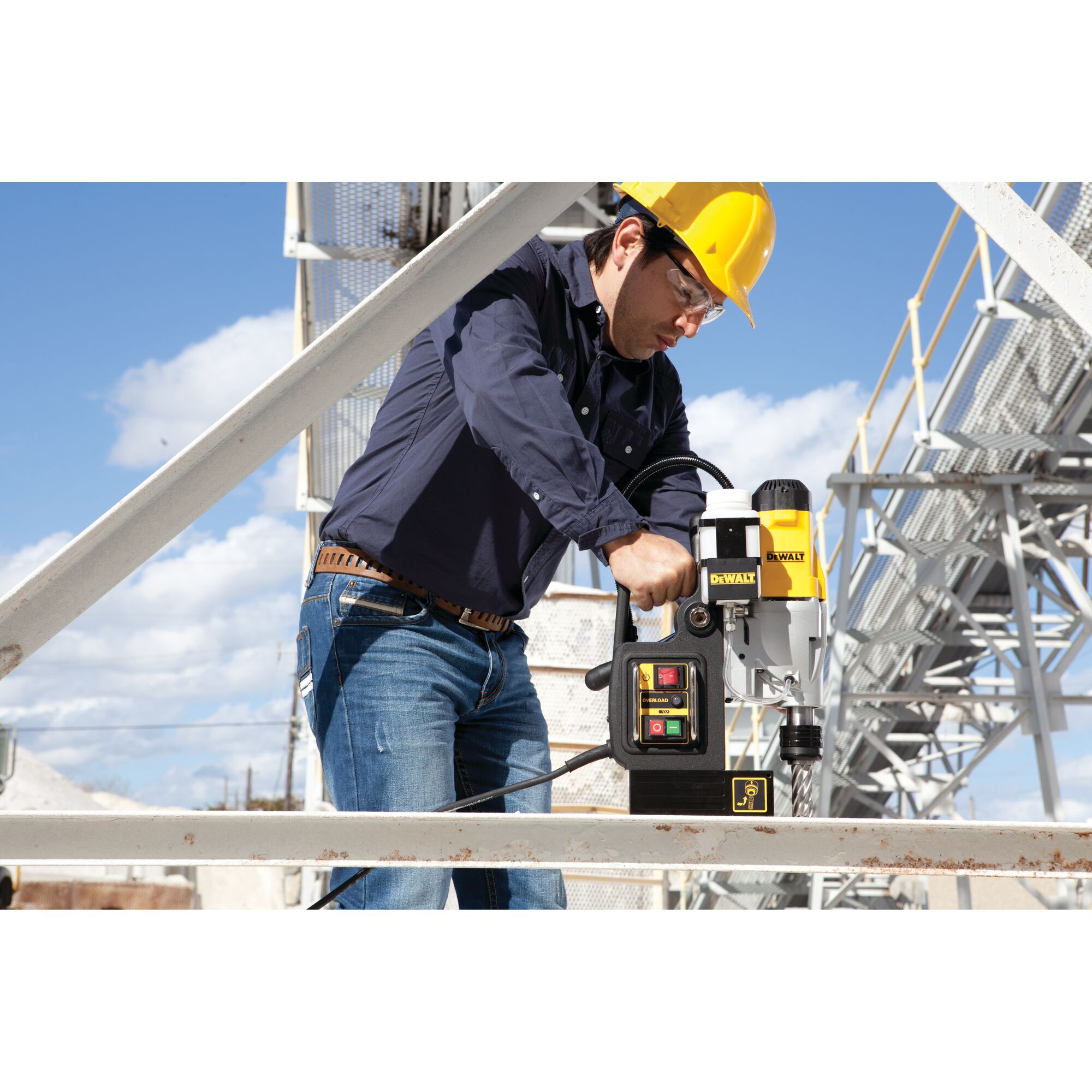 A person wearing a yellow safety helmet and blue jacket is using a DEWALT DWE1622K magnetic drill press on a metal beam at an outdoor industrial site. The face is blurred for privacy.