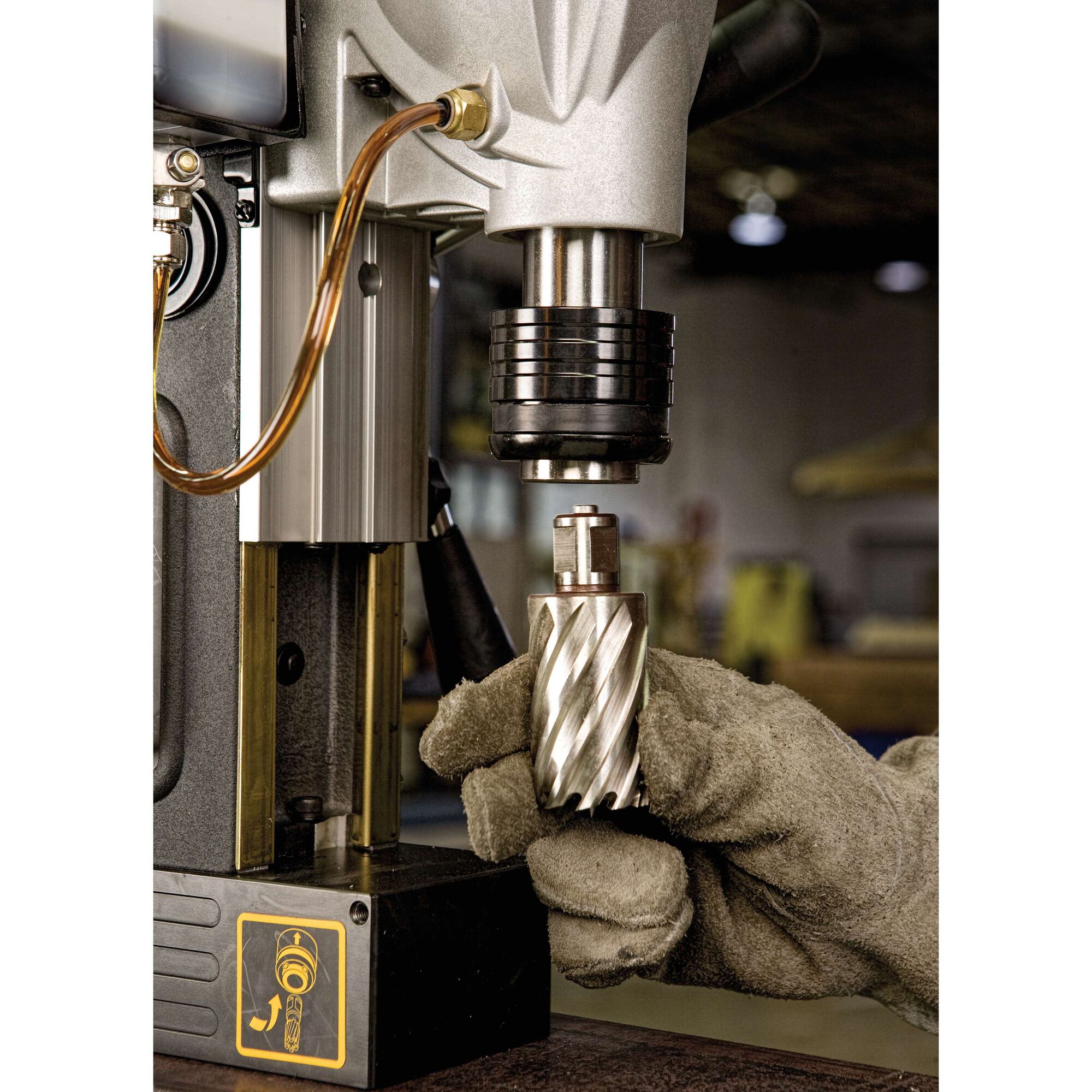 A close-up image of a worker's gloved hand inserting a metal drill bit into a DEWALT magnetic drill press. The scene is set in a workshop.