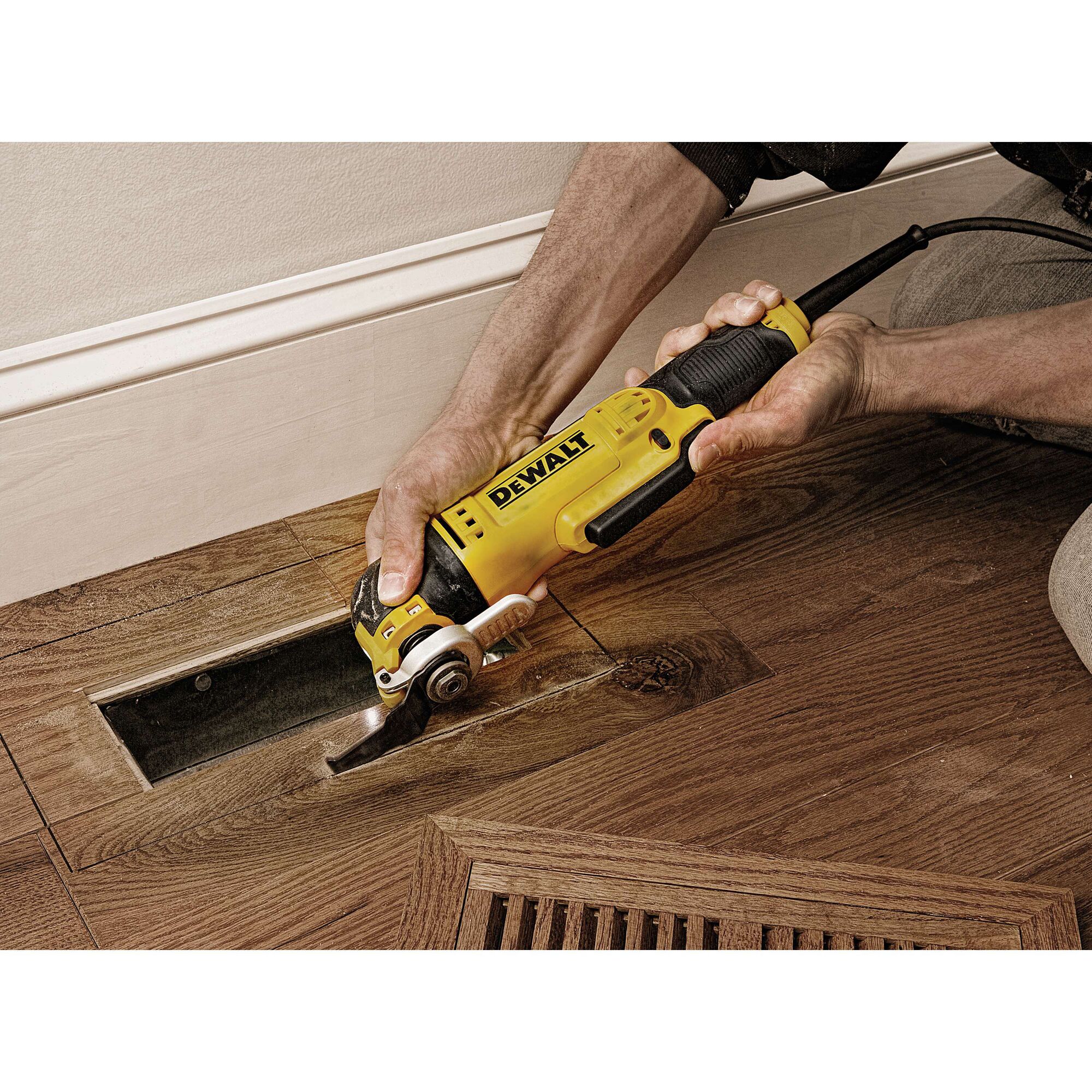 A person using a DEWALT DWE315 oscillating multi-tool to cut a section of hardwood floor near a wall.