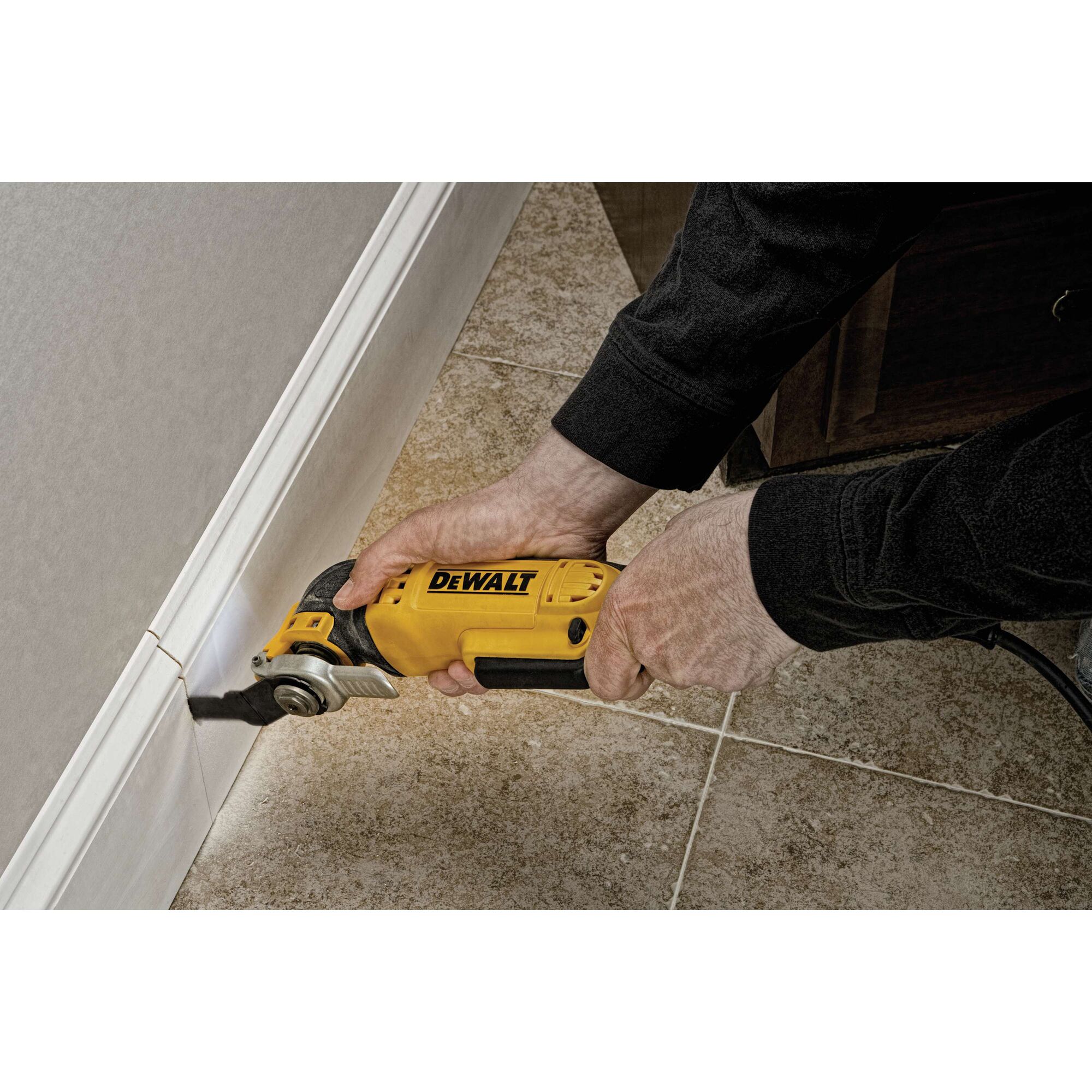 A person using a DEWALT oscillating multi-tool to cut along the baseboard edge of a tiled floor.