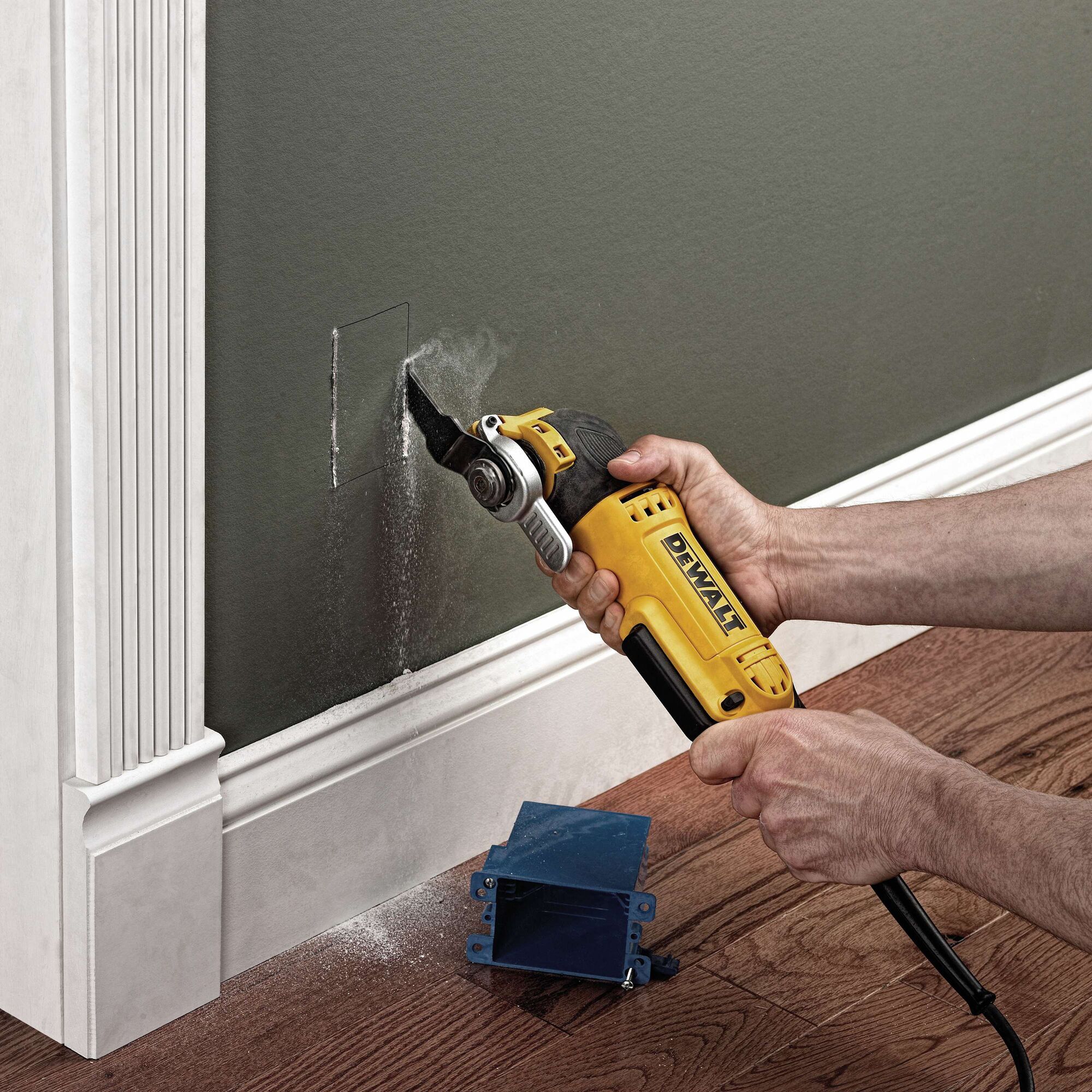 A person is using a yellow DEWALT DWE315 oscillating multi-tool to cut a rectangular hole in a wall near the floor. There is sawdust falling, and a blue plastic electrical box is placed on the wooden floor below.