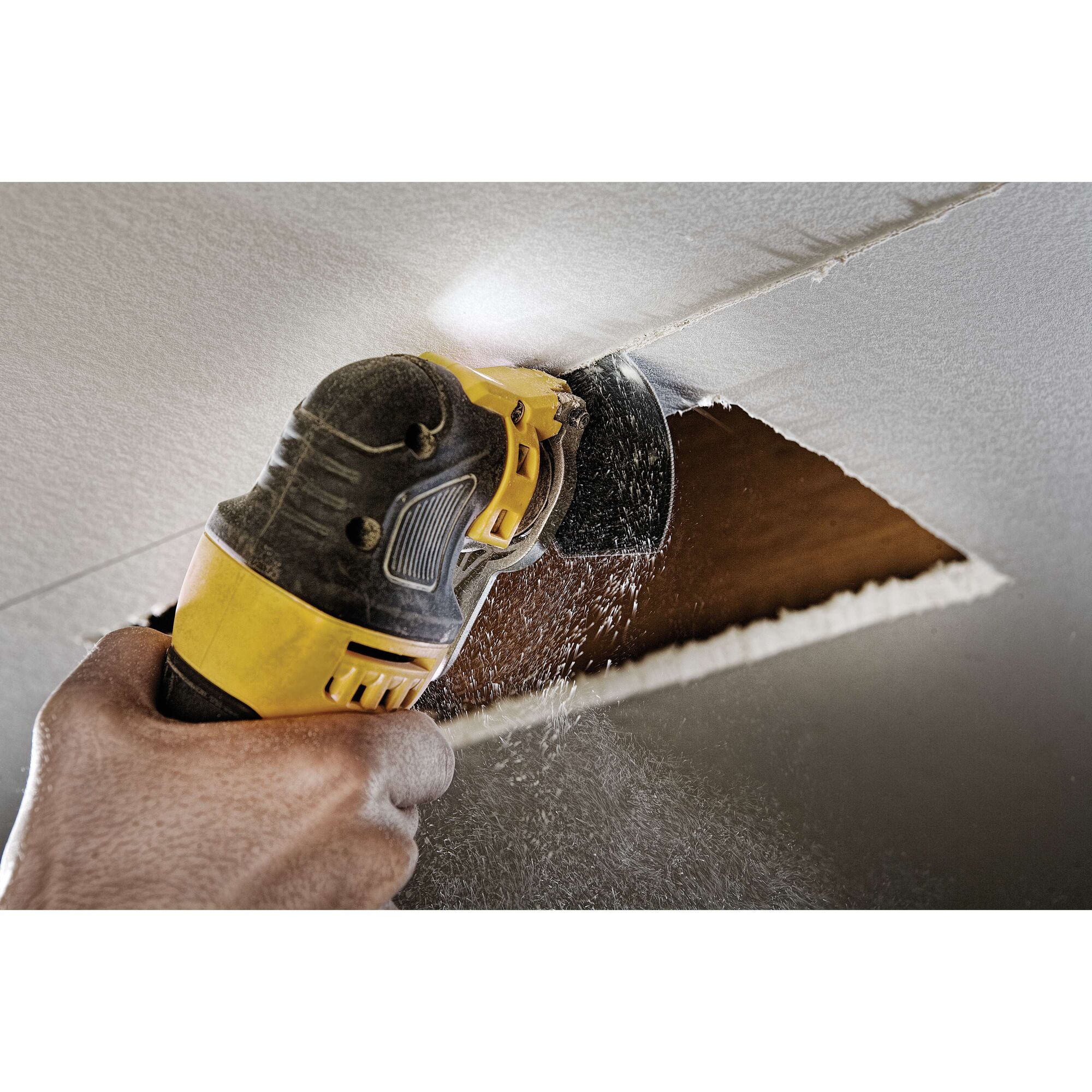 A close-up of a hand using a DEWALT oscillating multi-tool to cut a rectangular hole in a white drywall ceiling, with dust particles visible.