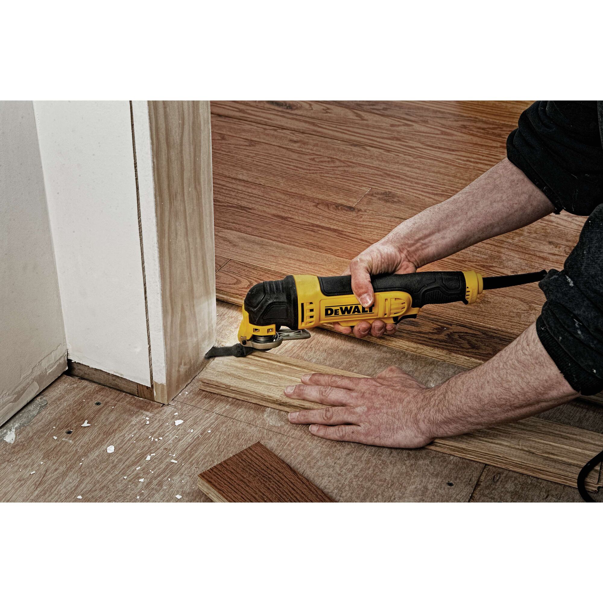 A person using a yellow and black DEWALT oscillating multi-tool to trim wood flooring near a door frame.