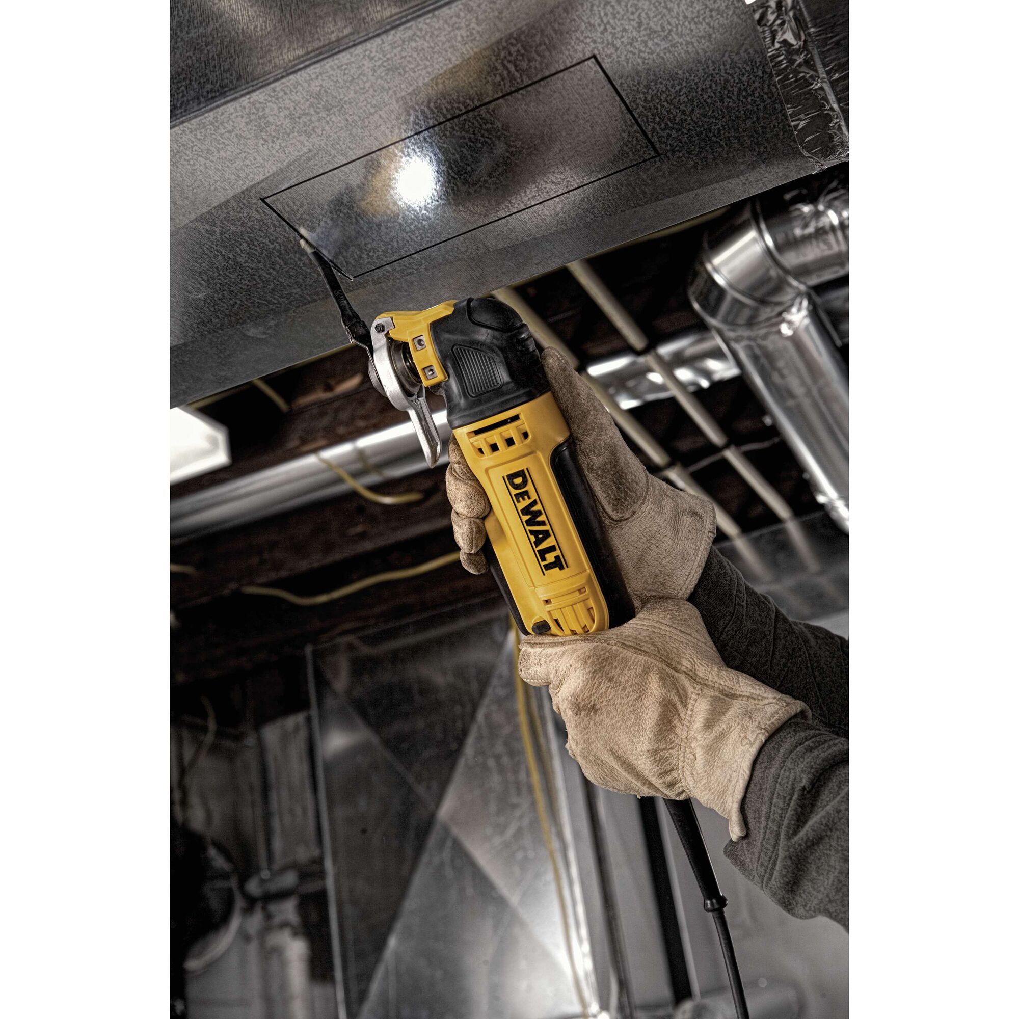 A person wearing work gloves uses a DEWALT oscillating multi-tool to cut a rectangular shape into a metal duct in an industrial setting.