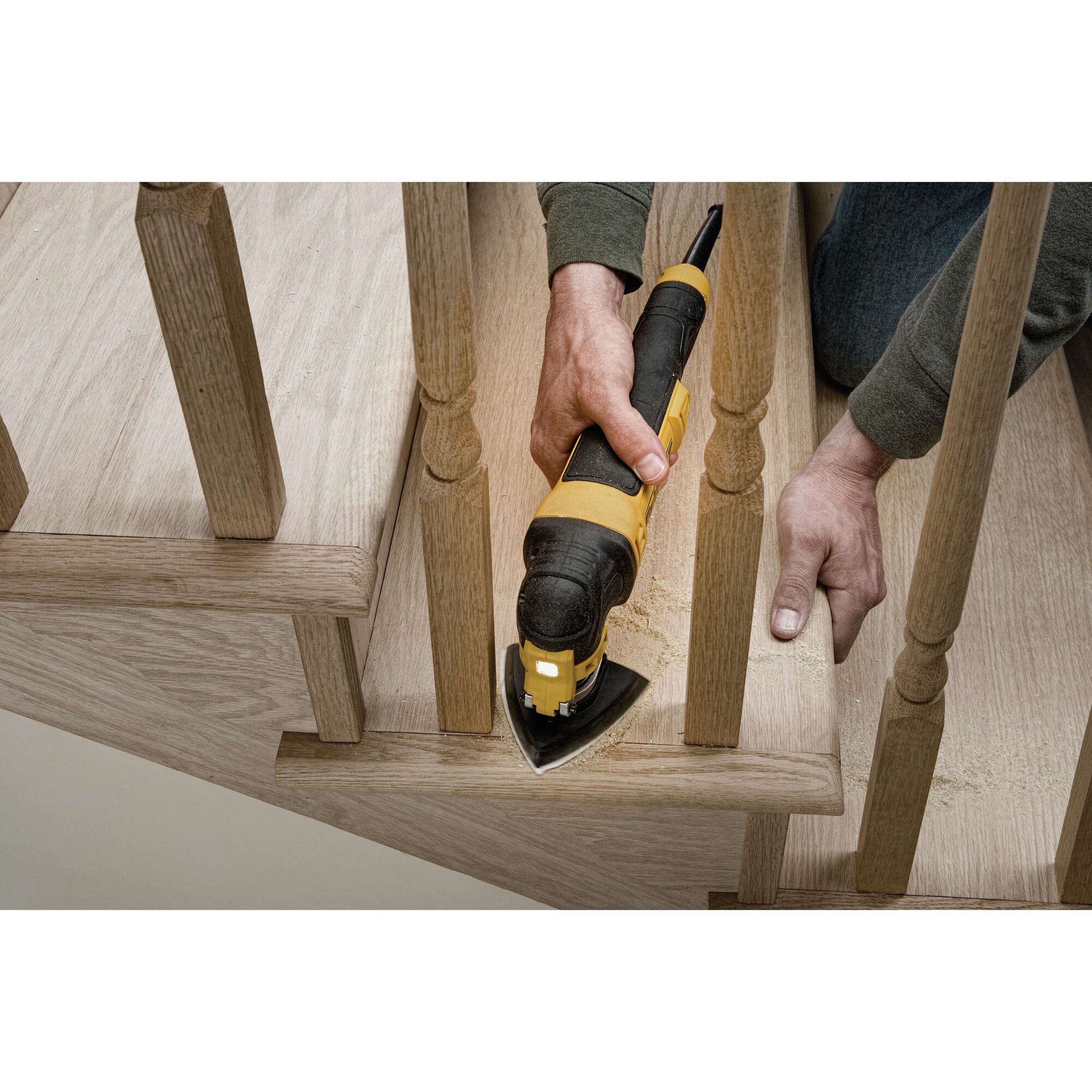 A person uses a DEWALT DWE315 oscillating multi-tool to sand or trim a wooden stair near the railing. The tool is held firmly and wood dust is visible.