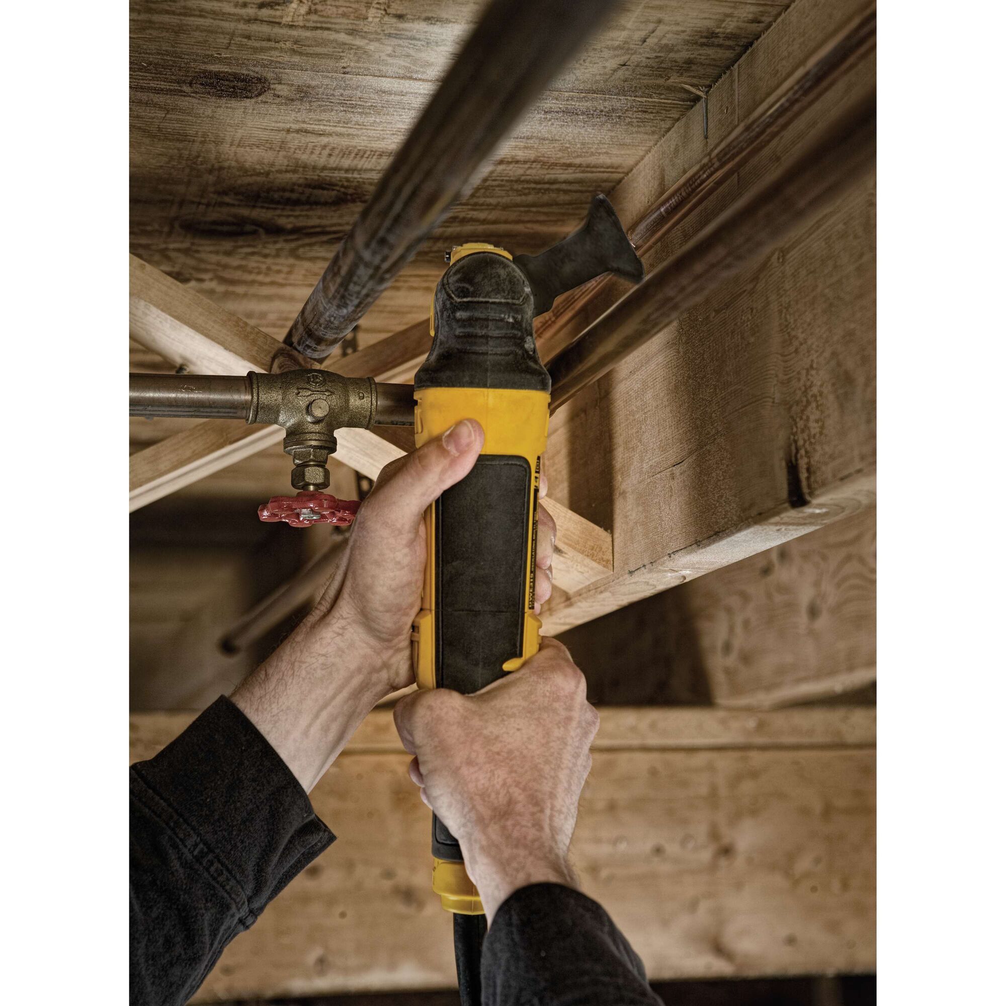 Hands using a DEWALT DWE315 oscillating multi-tool to cut a pipe under wooden beams.