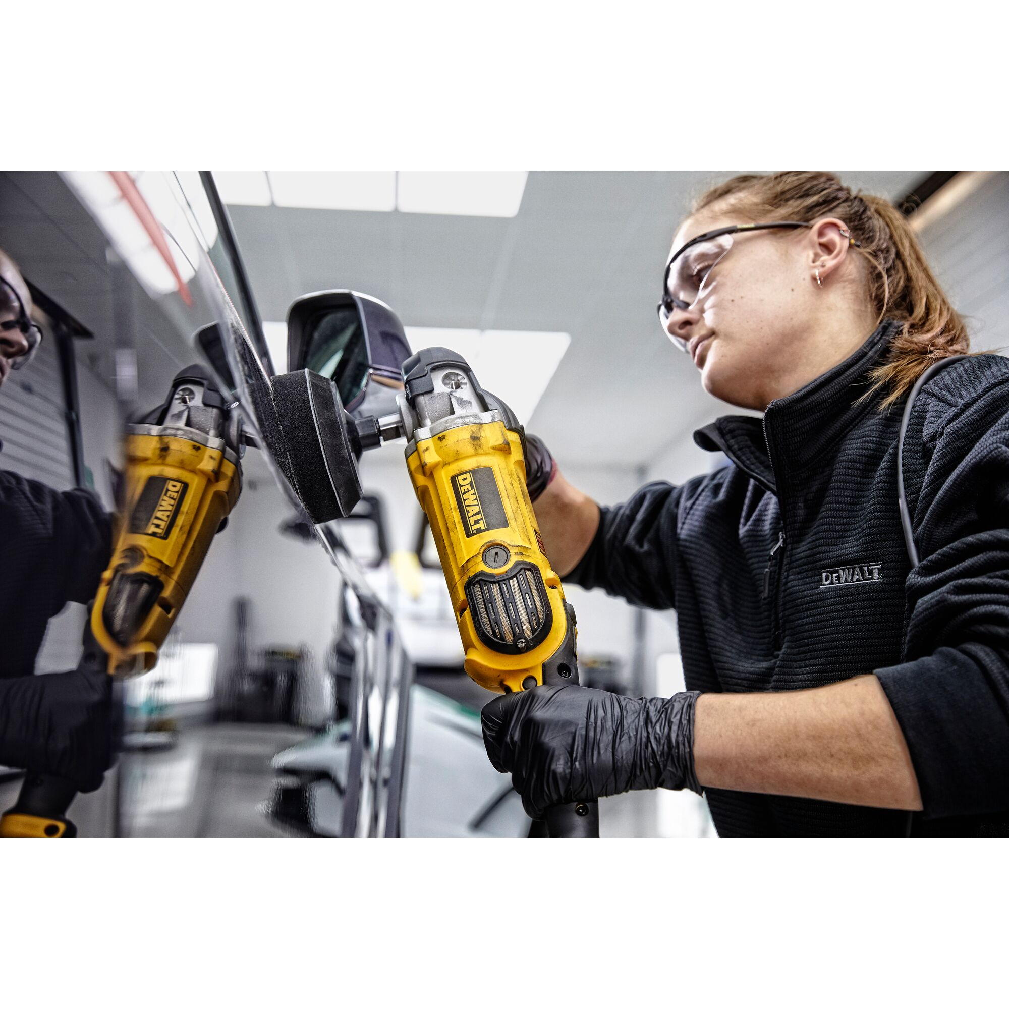 Person wearing black gloves and a DeWalt jacket using a yellow DeWalt polishing tool on the surface of a vehicle inside a workshop.