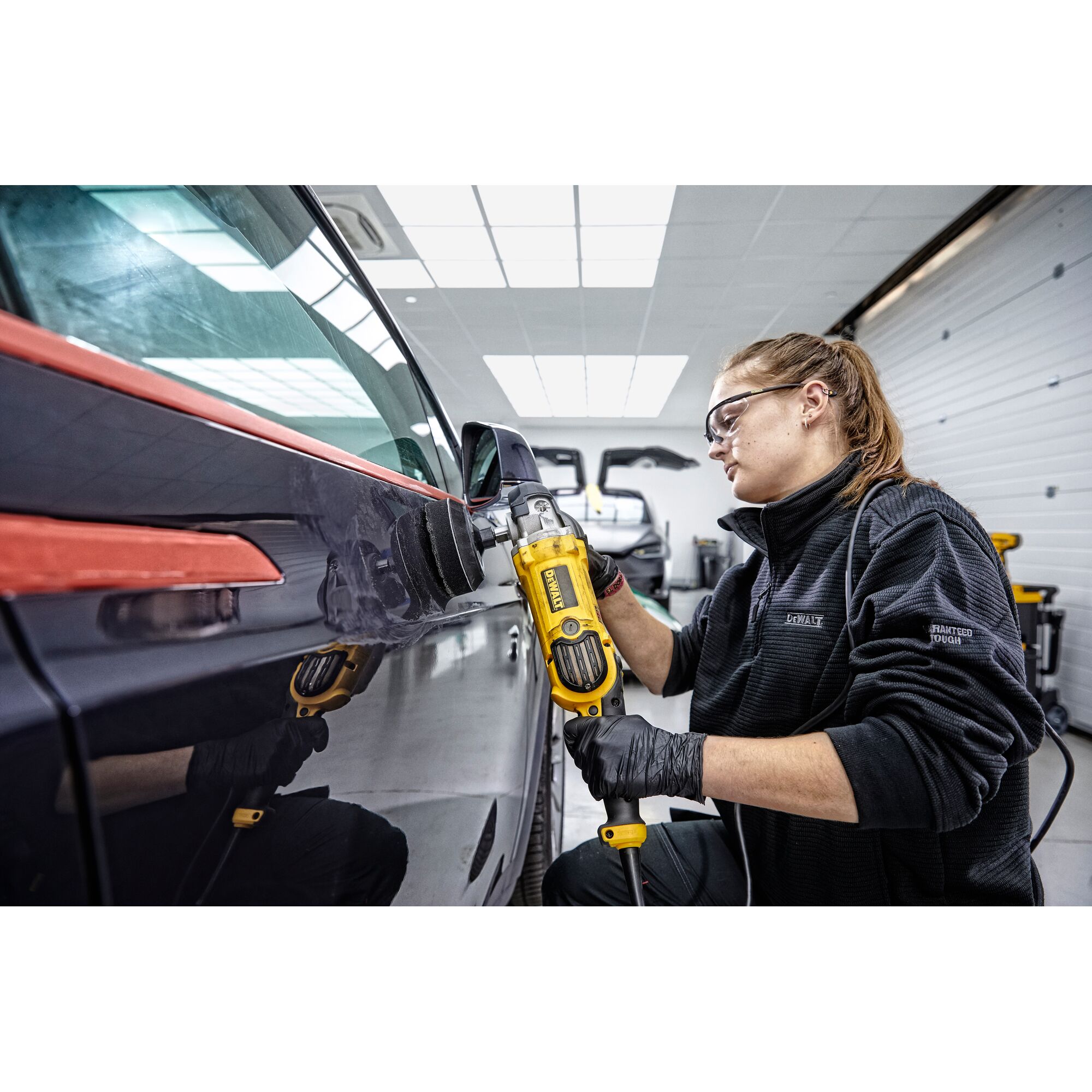 Person using a DEWALT DWP849X polisher on a car surface in a workshop. The individual is wearing black gloves and a dark jacket, and the setting is a brightly lit garage.