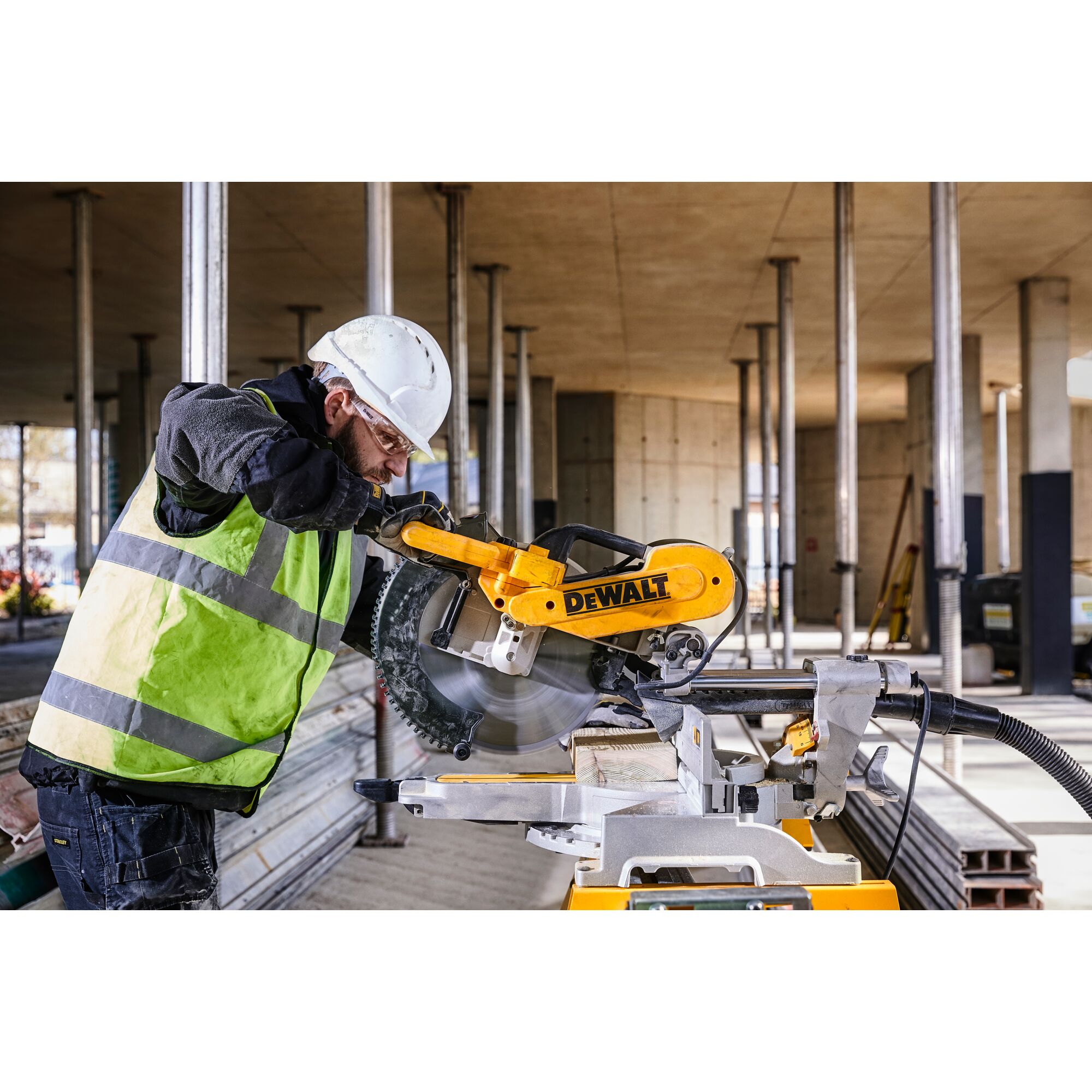 A construction worker wearing a safety vest and helmet operates a DEWALT DWS780 miter saw on a construction site. The worker's face is blurred for privacy.