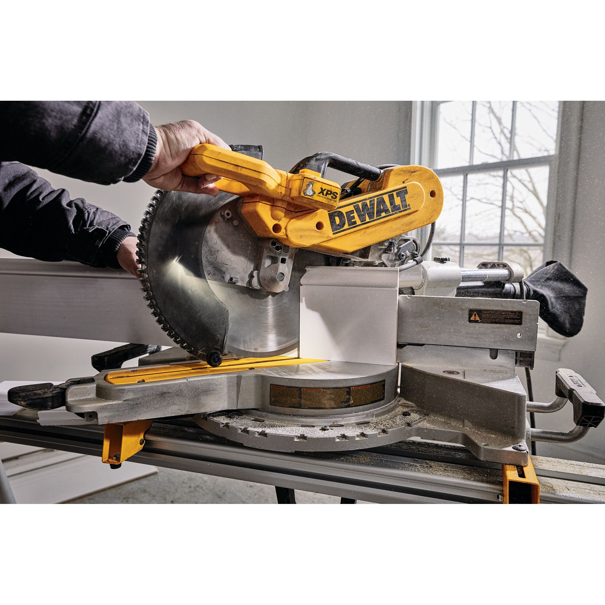 A close-up view of a DEWALT DWS780 miter saw being used by a person to cut a piece of wood indoors. The saw is mounted on a workbench, with natural light coming in from a window in the background.