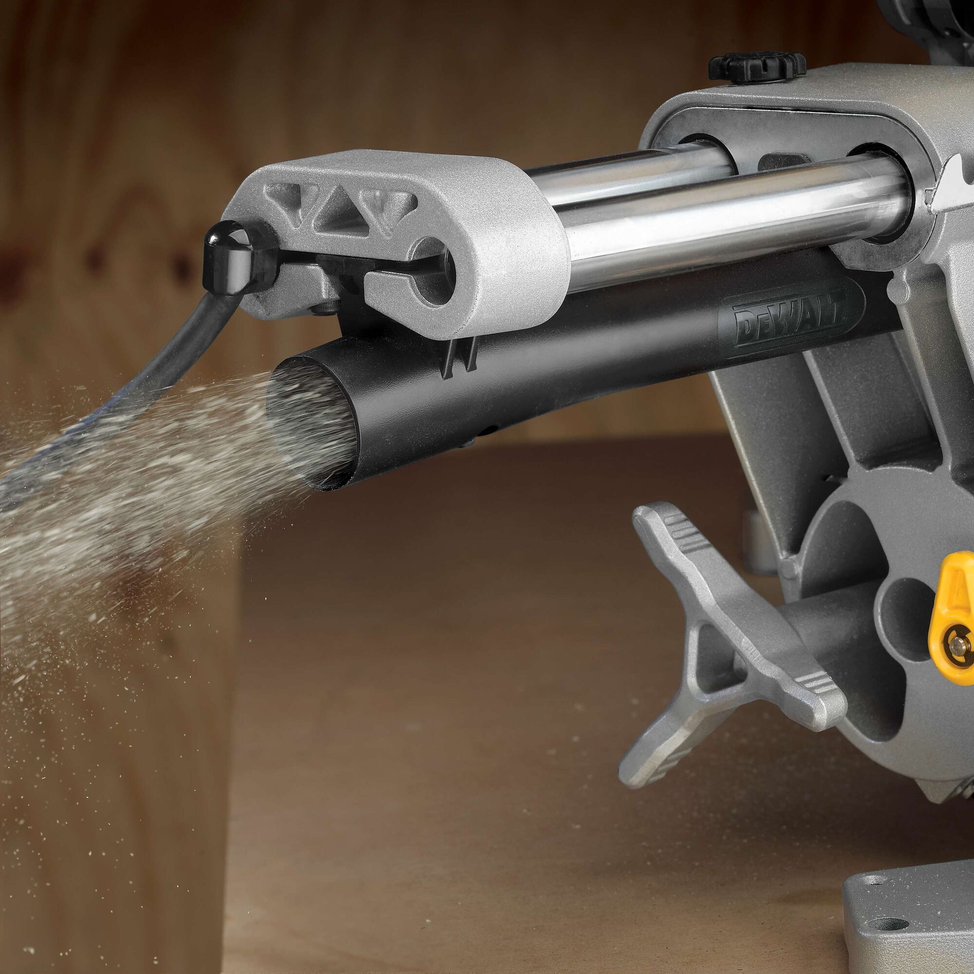 Close-up view of the dust collection system of a DEWALT miter saw, showing dust being expelled through a black tube attached to the saw.